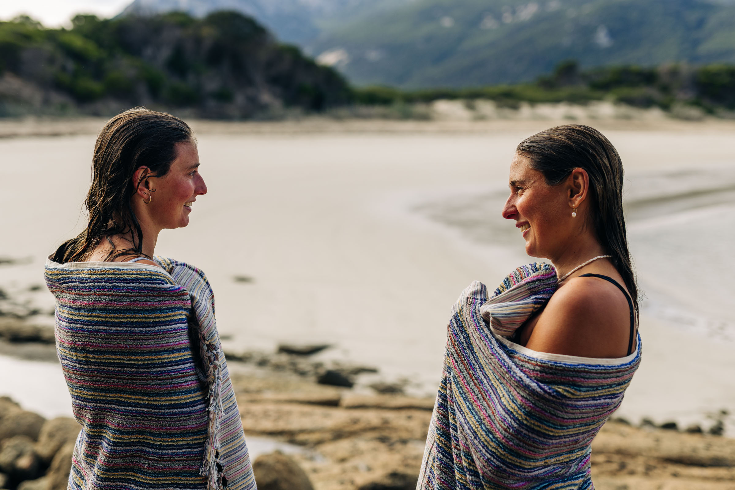 Two women wrapped in striped blankets smiling at each other on a rocky beach at sunset.