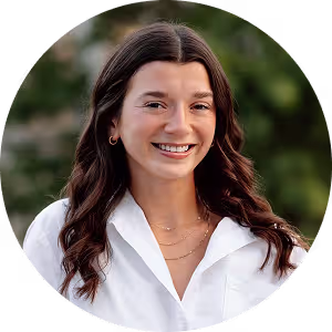 Smiling woman with long dark wavy hair wearing a white blouse and gold jewelry outdoors.