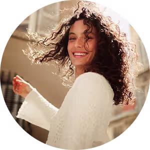 Smiling woman with curly hair wearing a white sweater, turning outdoors in sunlight.