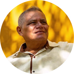 Smiling older man wearing a light shirt against a bright yellow background with sunlight shadows.