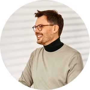 Smiling man with glasses and short dark hair wearing a beige sweater and black turtleneck.