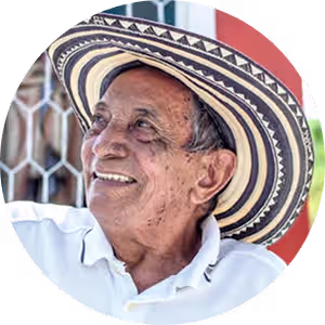 Smiling older man wearing a traditional wide-brimmed hat and white shirt, looking to the side.
