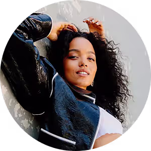 Confident young woman with curly hair in black leather jacket reclining against wall.