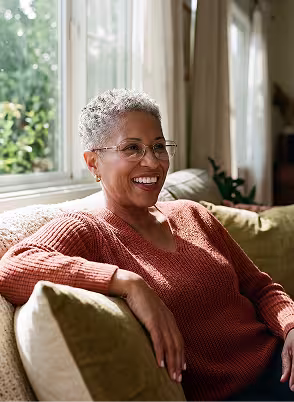 Smiling elderly woman with glasses wearing a rust-colored sweater sitting on a couch by a window.