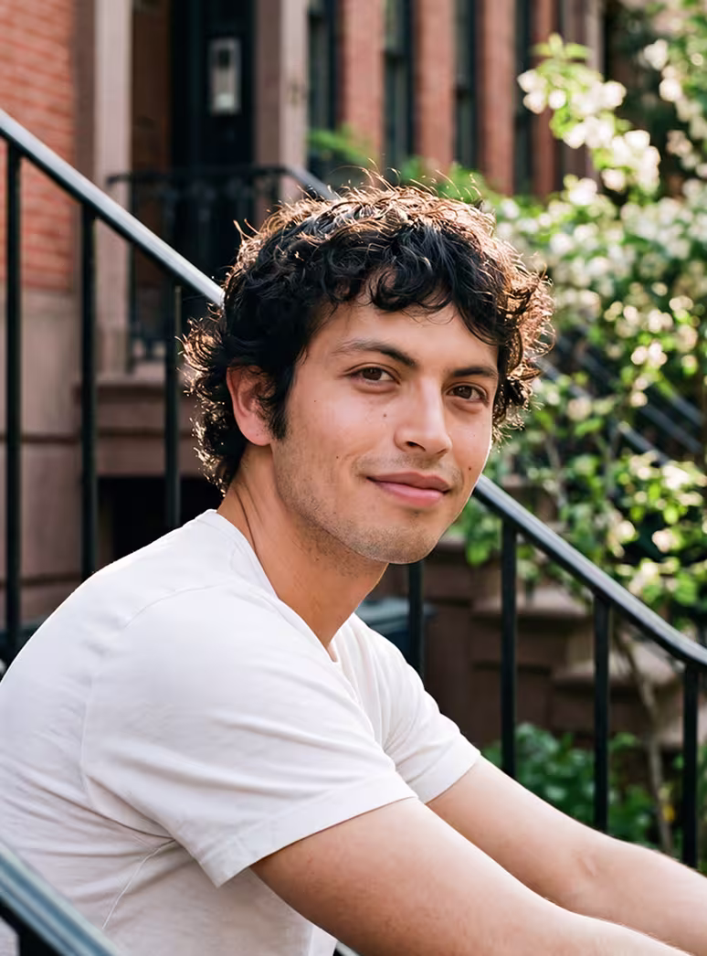 Young man with curly hair wearing a white t-shirt sitting outdoors by a black railing and brick building.