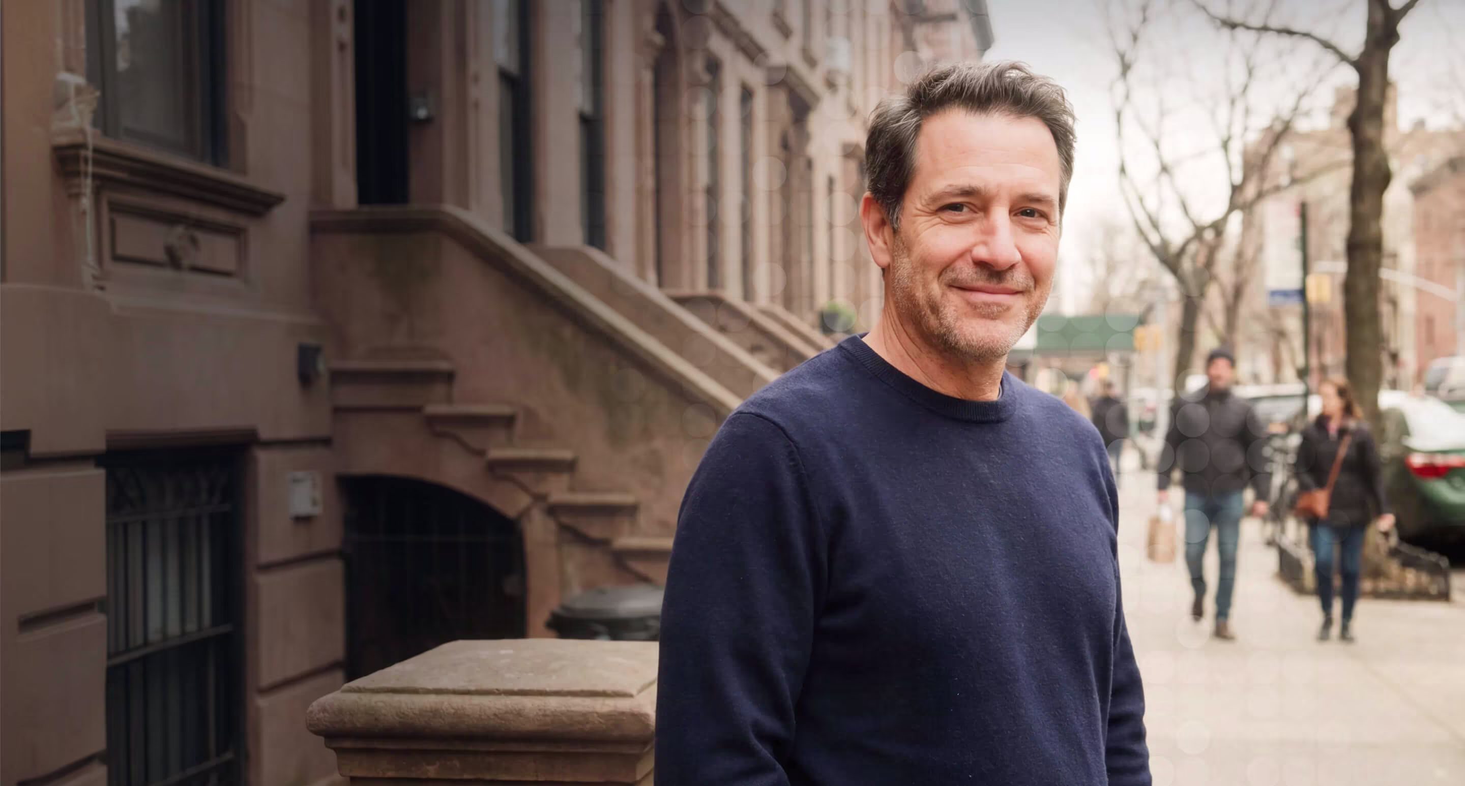 Smiling man in navy sweater standing on sidewalk by stone stairs and brownstone buildings.