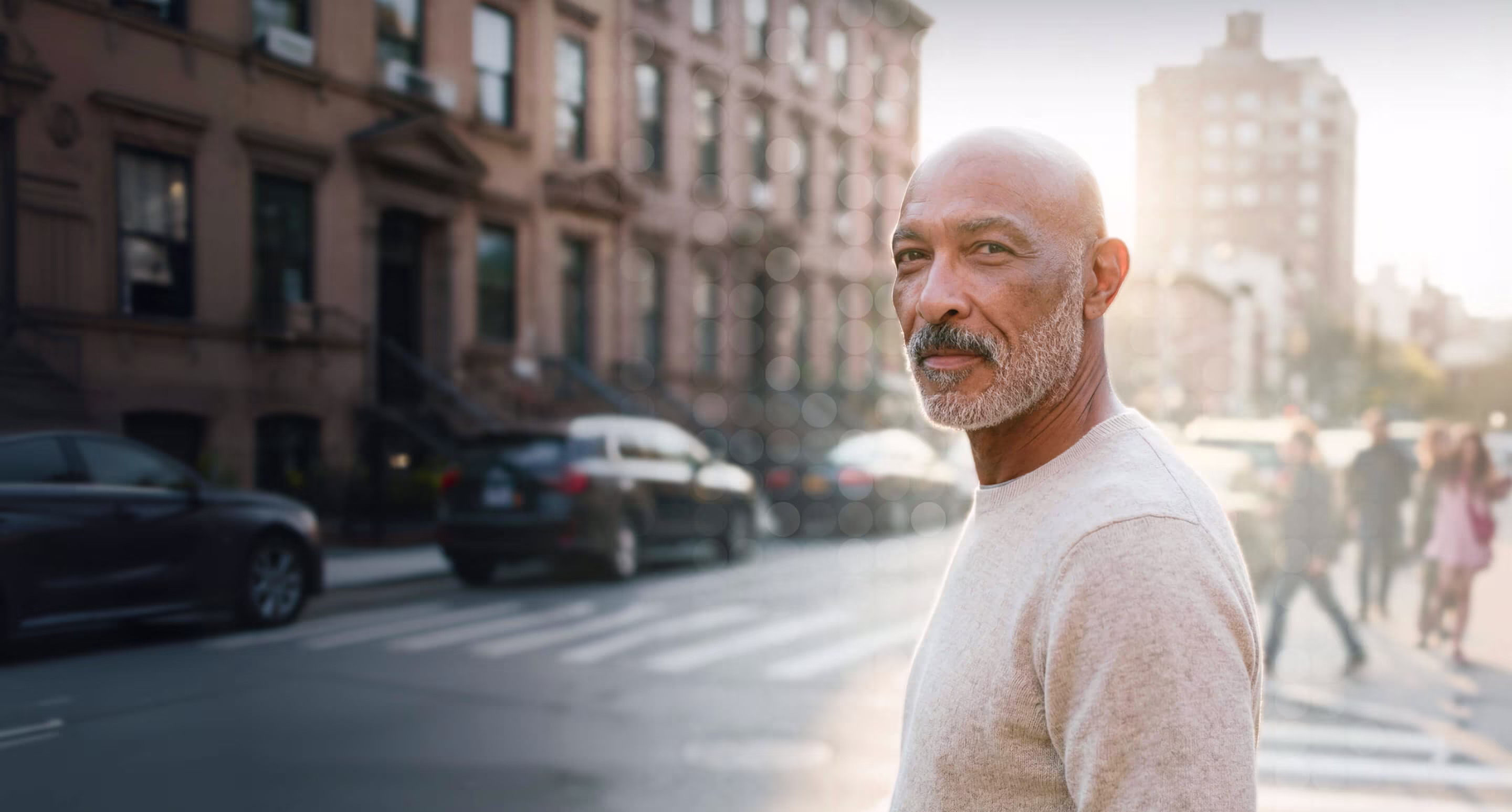 Bald man with gray beard wearing light sweater standing near street with blurred buildings and cars.