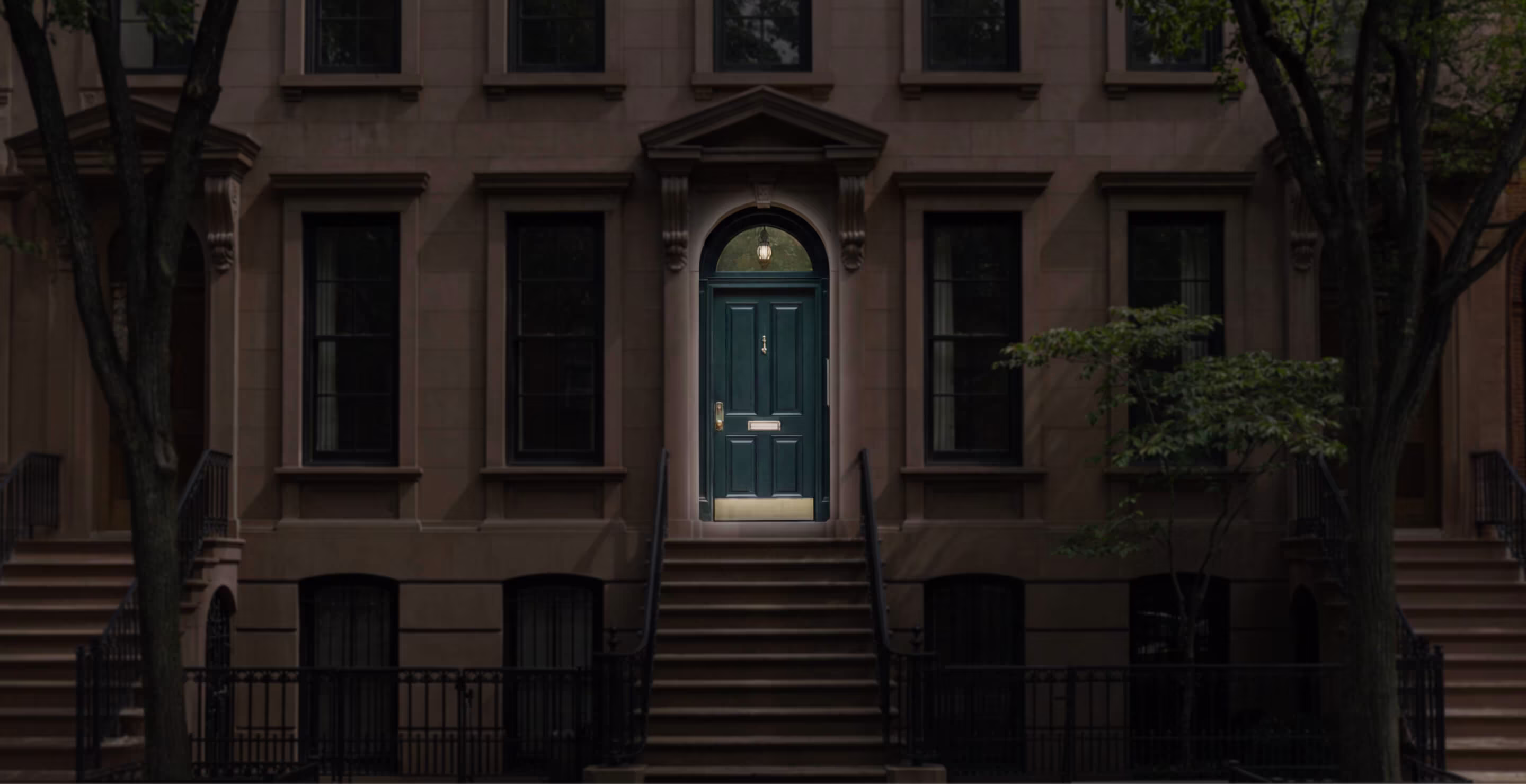Dark green door illuminated by light in brownstone building with stairs and trees in front.