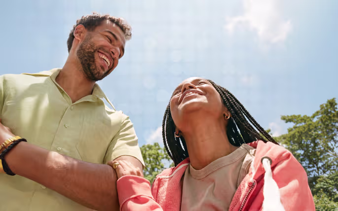 Man and woman smiling and holding hands outdoors under a clear blue sky with trees.