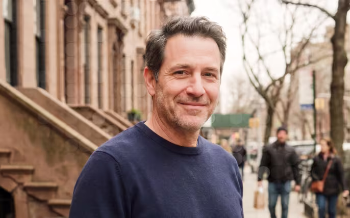 Smiling middle-aged man with dark hair wearing a navy sweater on a city street with brownstone buildings.