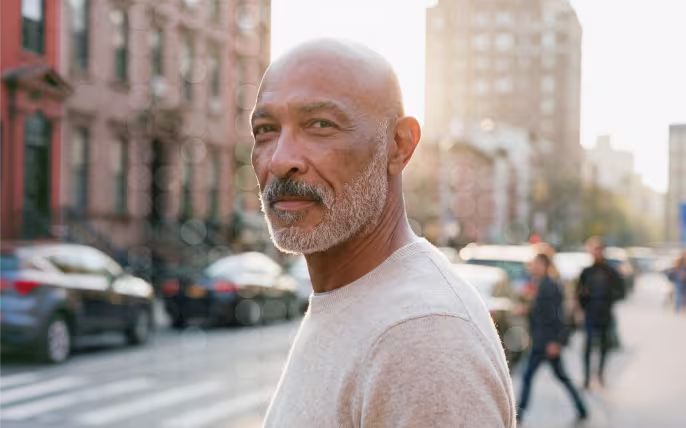 Bald man with gray beard wearing light sweater standing outdoors with blurred city street background.