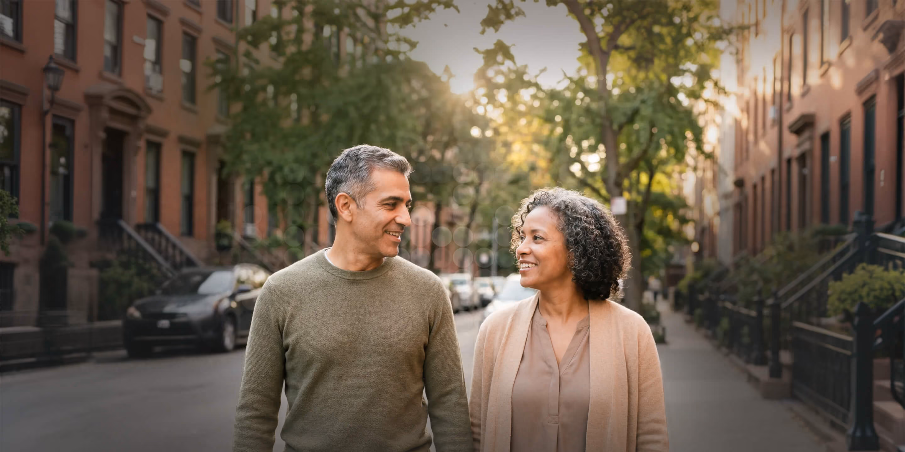 Middle-aged couple smiling and walking together on a tree-lined urban street with brownstone buildings.