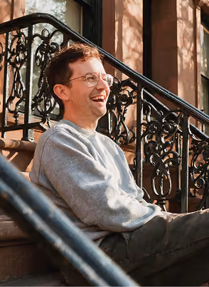 Young man with glasses laughing while sitting on outdoor stairs with ornate railing.