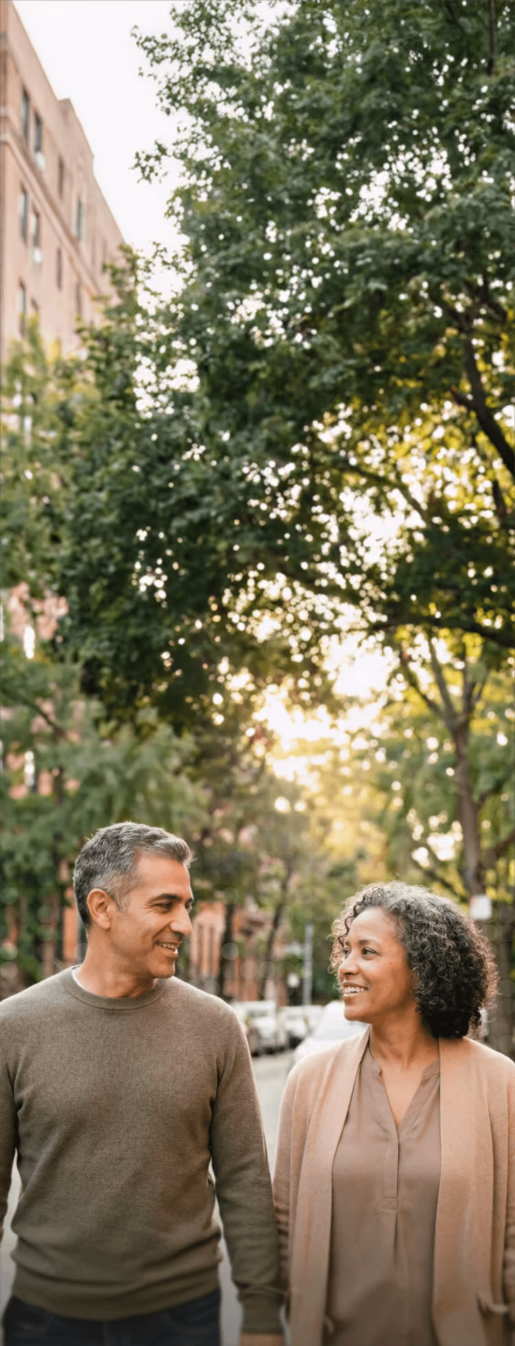 Middle-aged man and woman walking outside smiling at each other on a tree-lined street.