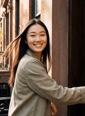 Smiling woman with long hair wearing a green jacket opening a brown door outdoors.