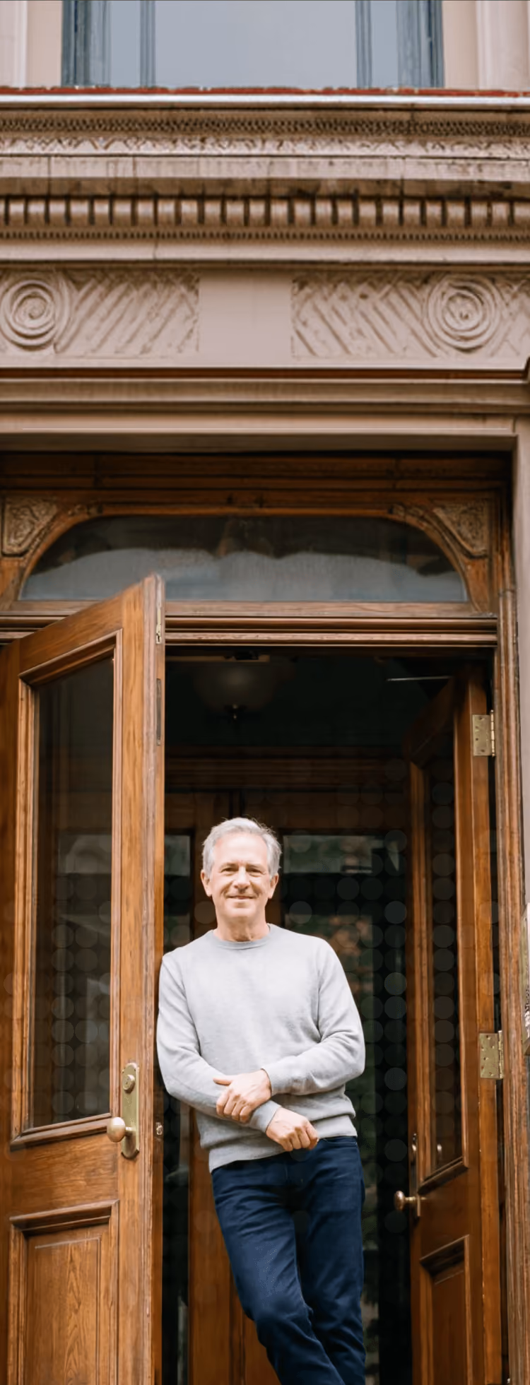 Smiling older man in gray sweater standing in the doorway of a building with wooden doors.