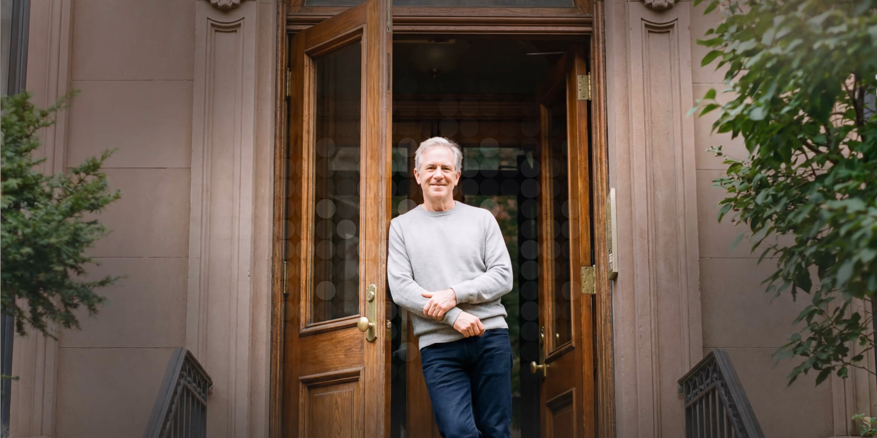 Smiling middle-aged man in a gray sweater standing in front of open wooden double doors of a building.