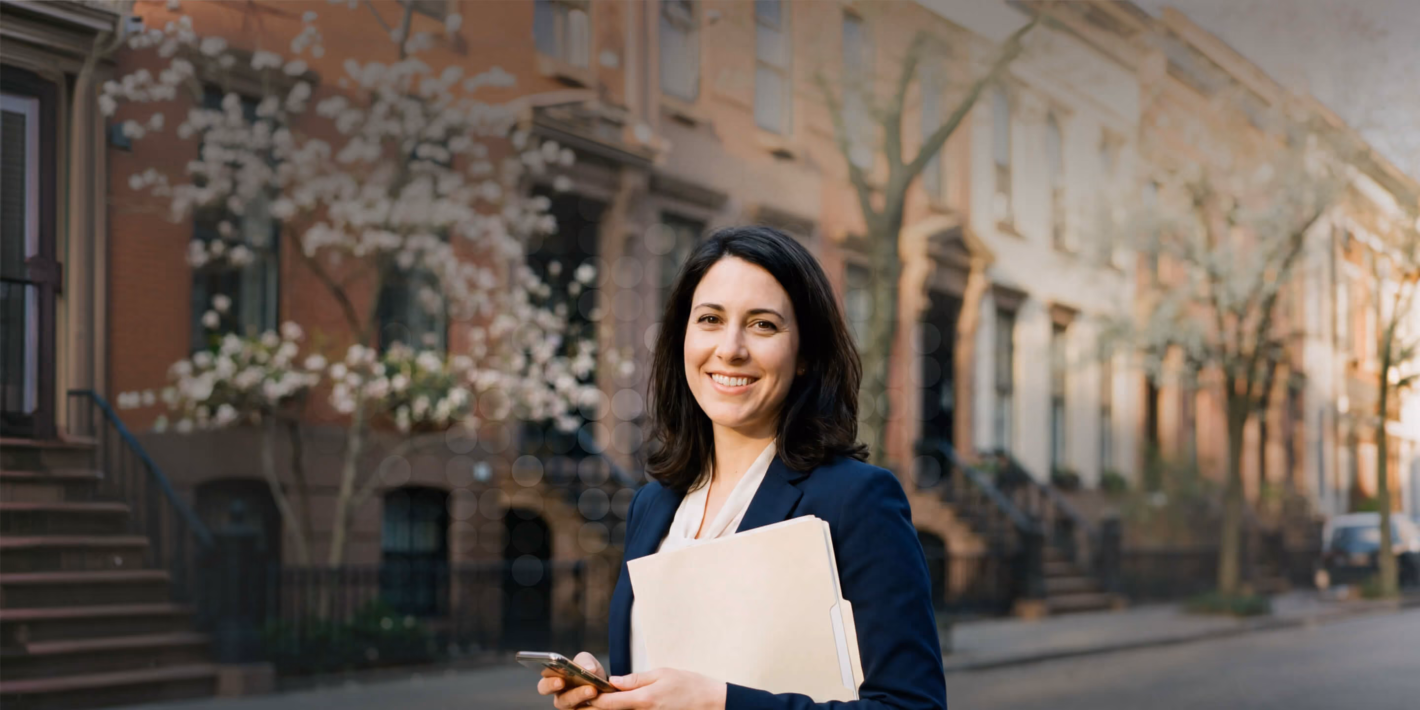 Smiling woman in blazer holding folder and phone on a city street with brownstone buildings.