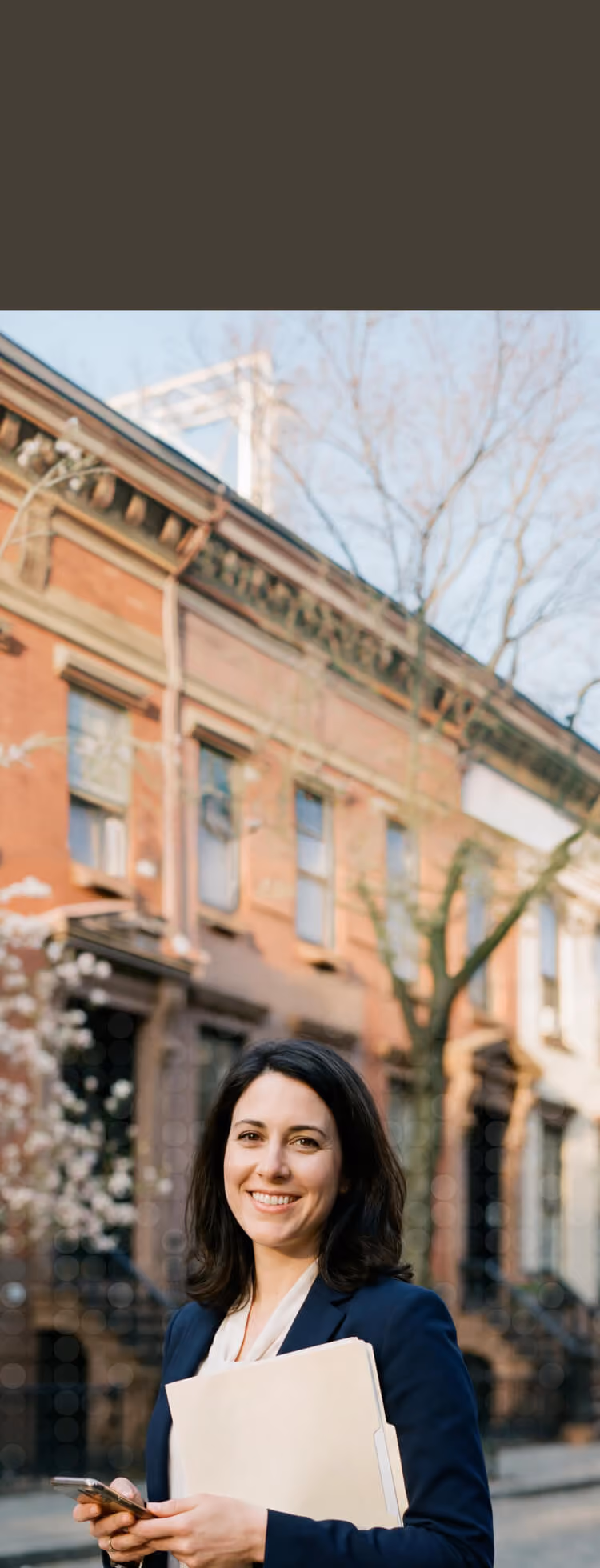 Smiling woman in business attire holding folder and phone on a city street with brownstones behind.