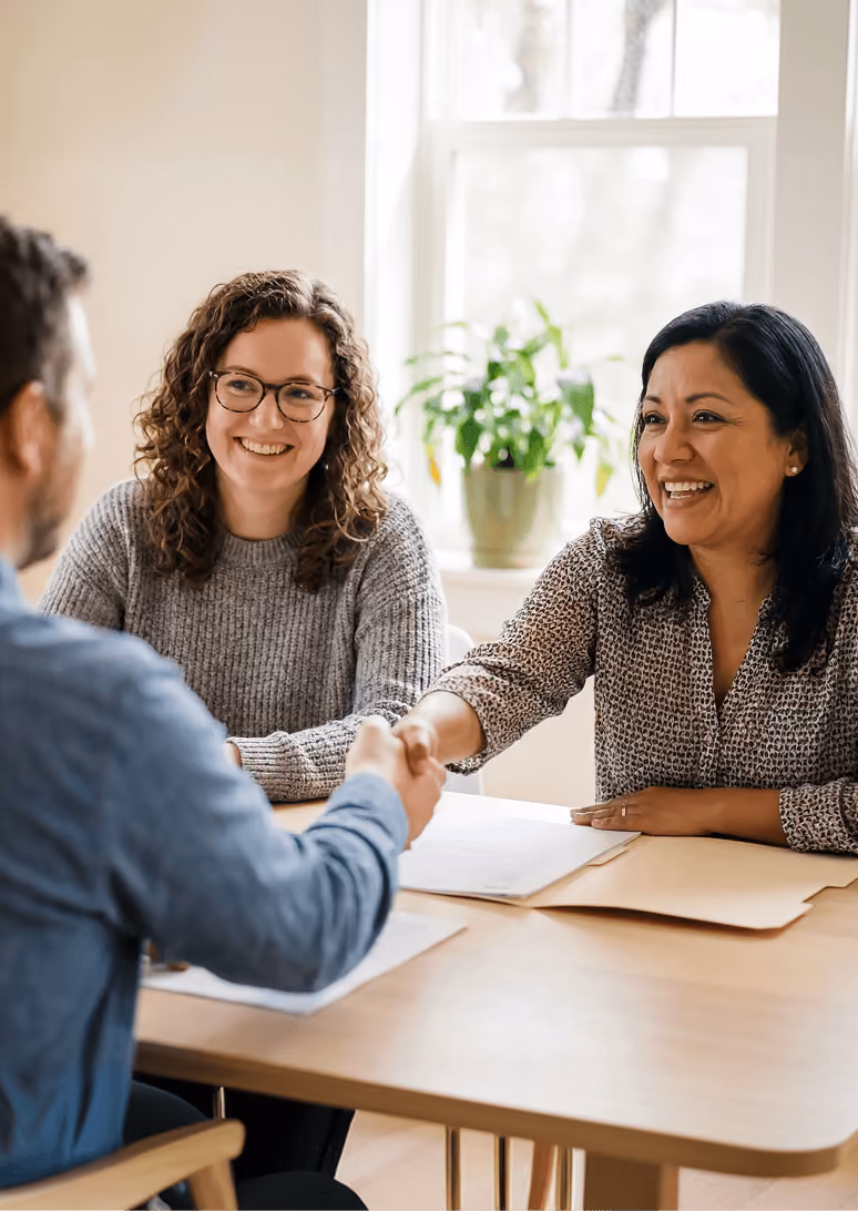 Two women smiling while shaking hands with a man across a table in a bright room.