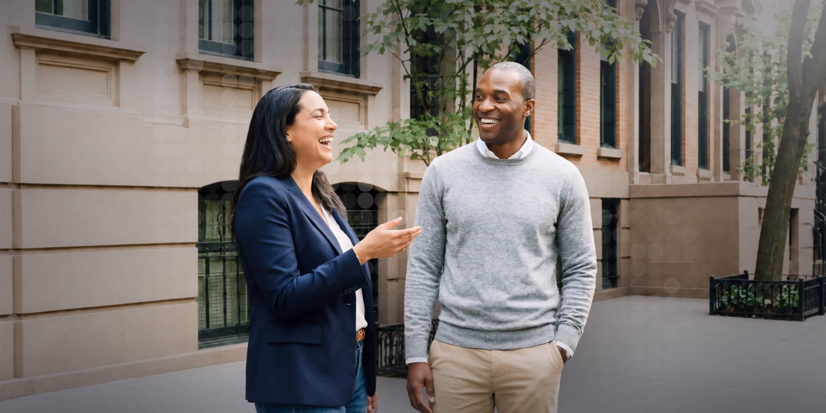 Man and woman smiling and talking outside near city buildings and trees.