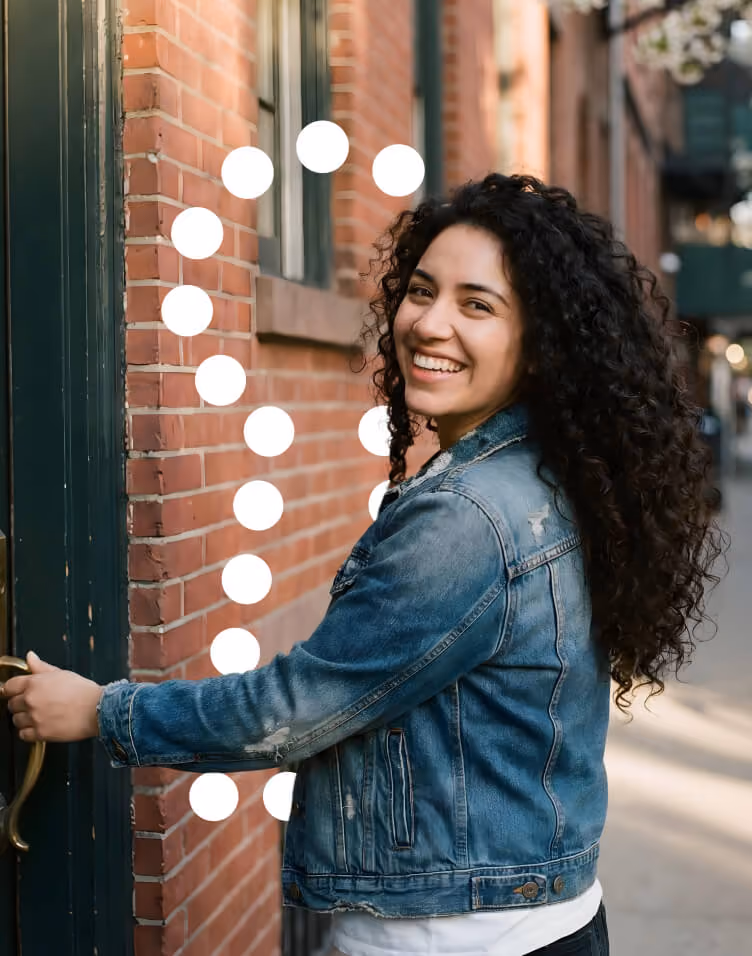 Smiling woman with curly hair wearing a denim jacket opening a door on a brick building.