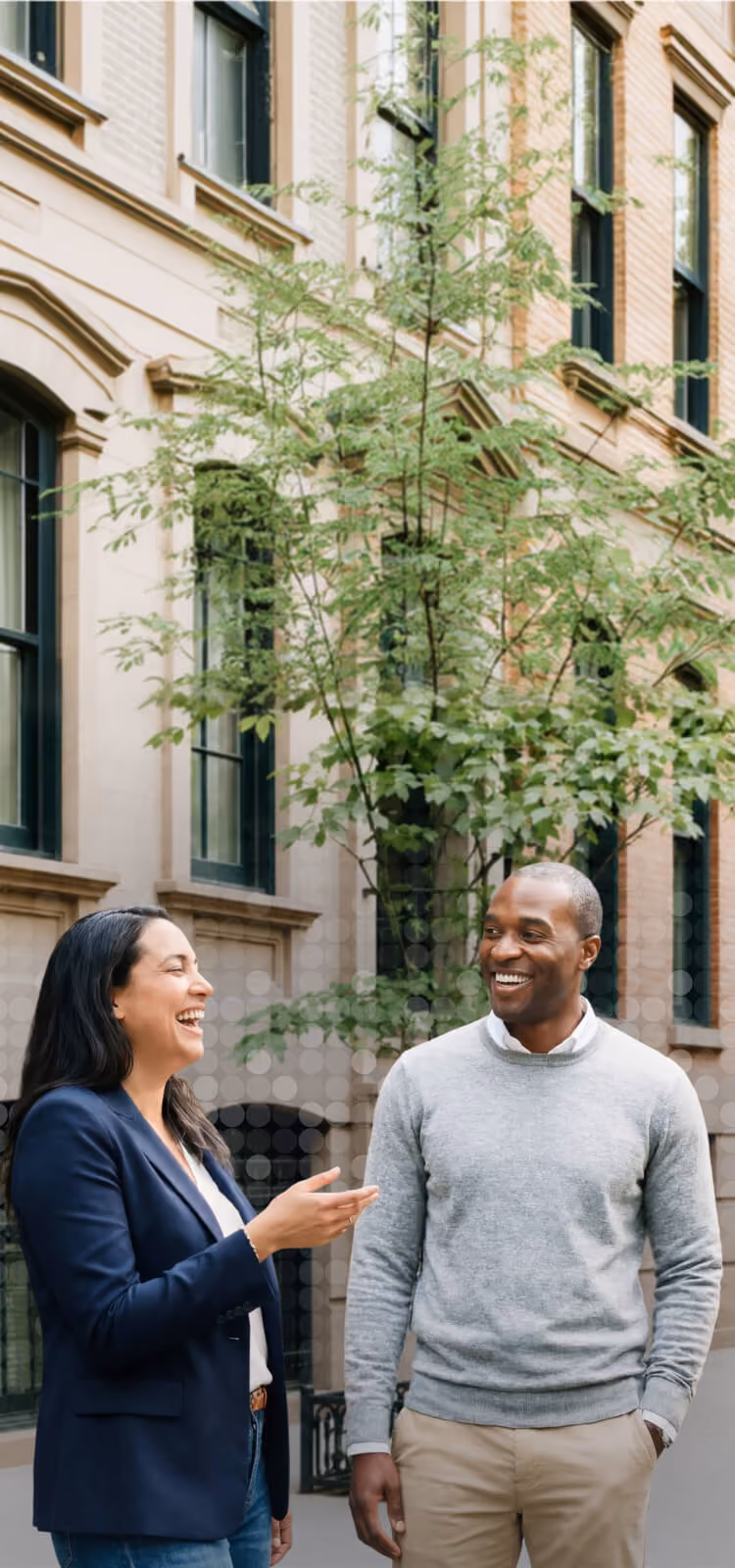 Man and woman smiling and talking outdoors in front of a brick building with green windows and trees.