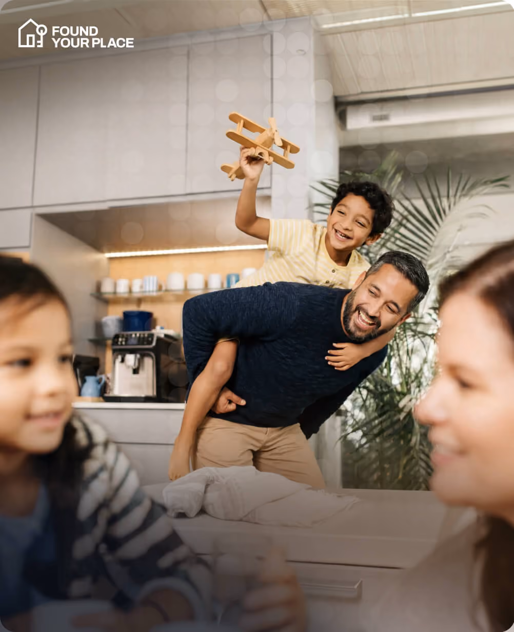 Man giving piggyback ride to smiling boy holding wooden toy airplane in a modern kitchen.