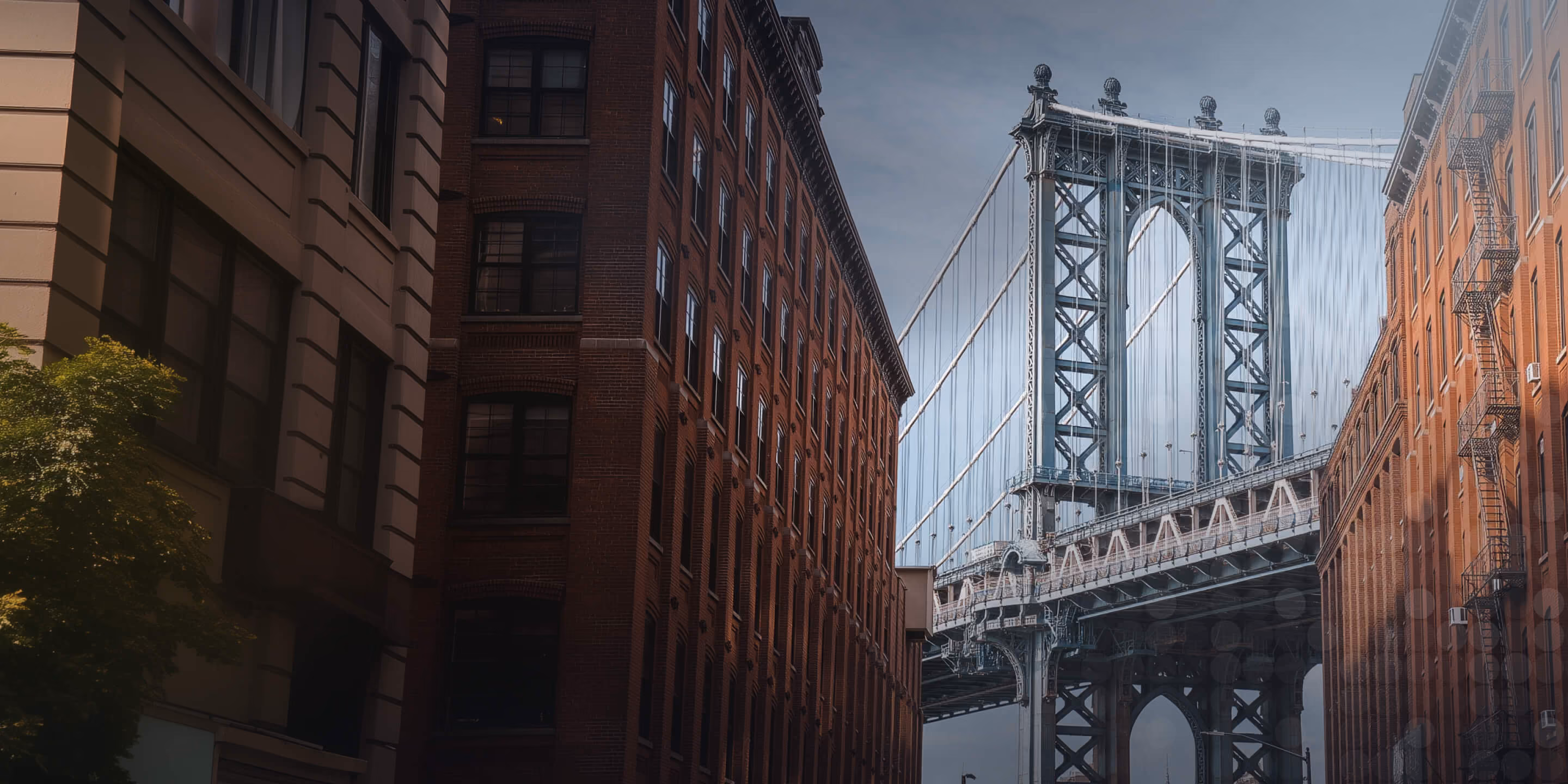 View of a large steel suspension bridge framed by brick and stone buildings on a cloudy day.