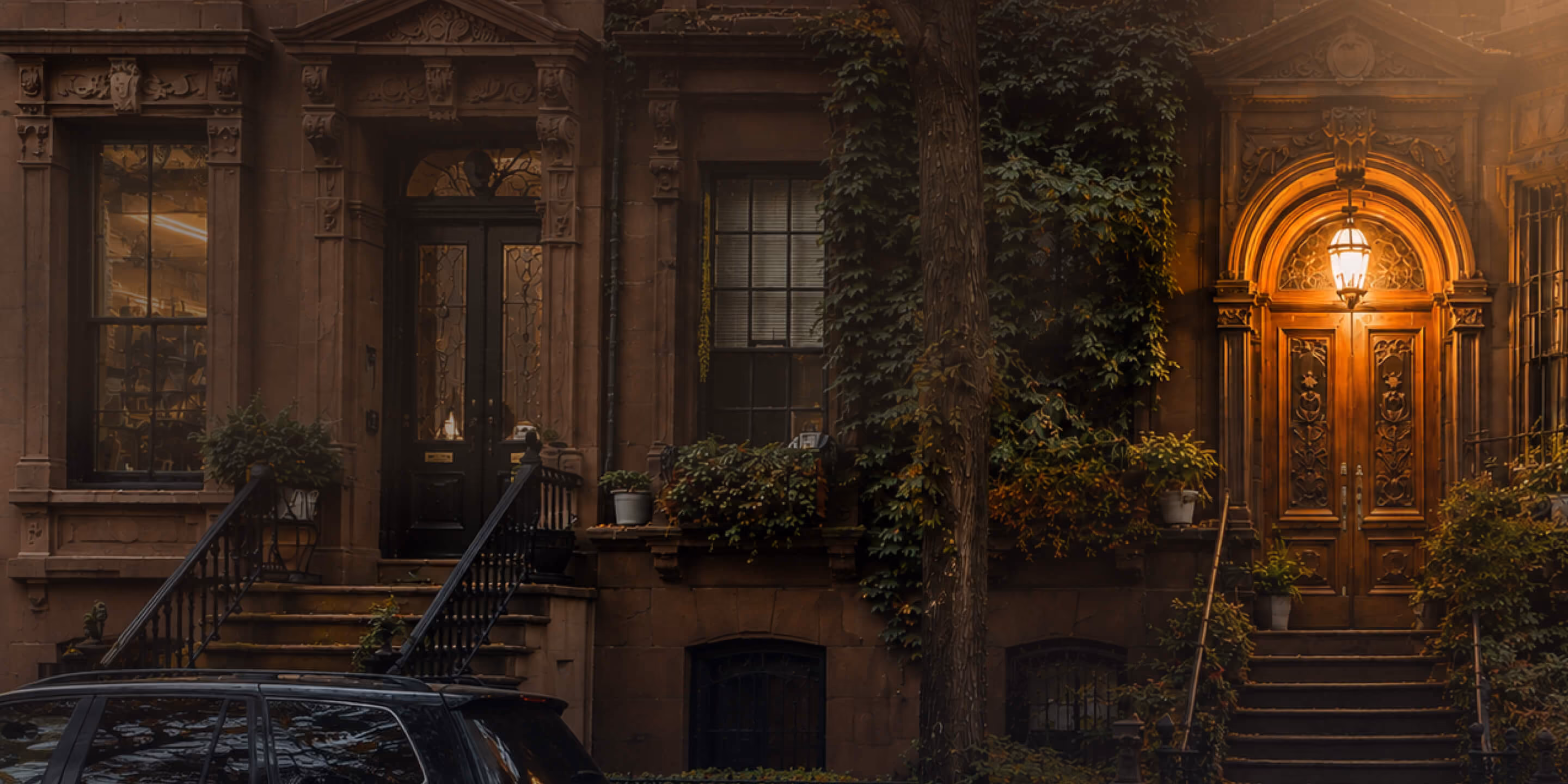 Ornate brownstone entrance with steps, plants, and a glowing lantern above a carved wooden door.