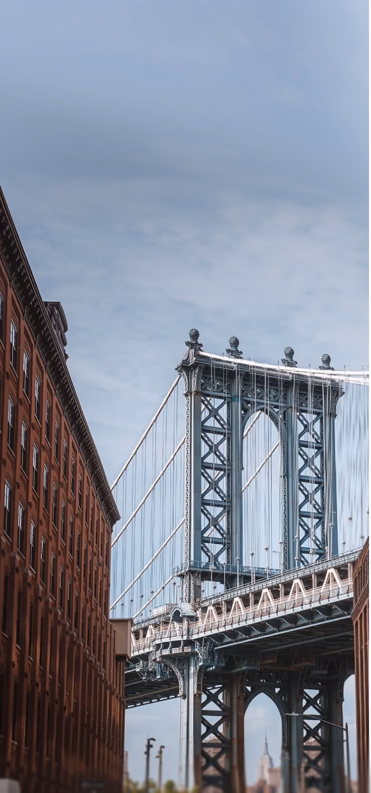 Steel suspension bridge tower with cables next to a tall brick building under a cloudy sky.