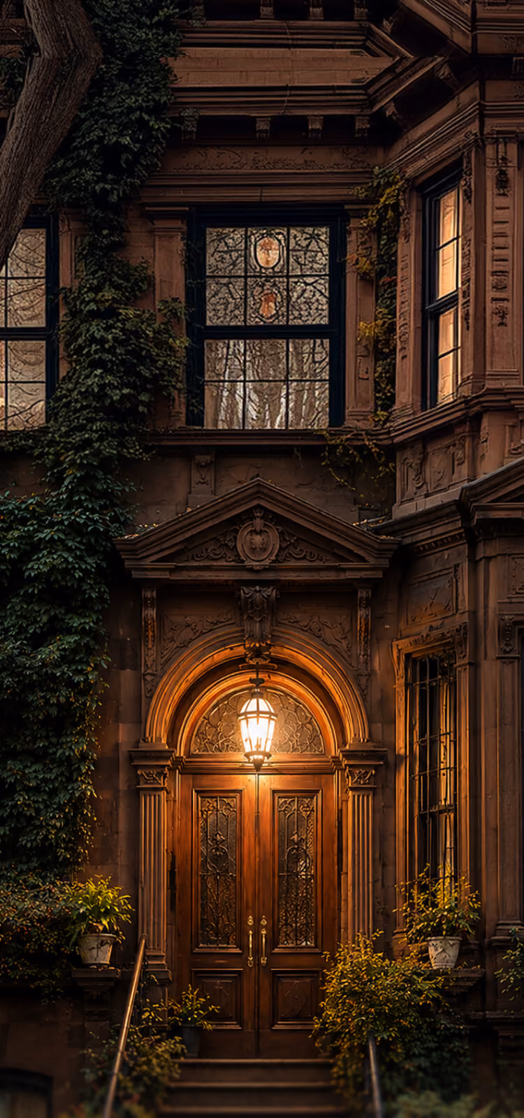 Ornate wooden door with stained glass and lit lantern framed by plants and stone architecture at dusk.
