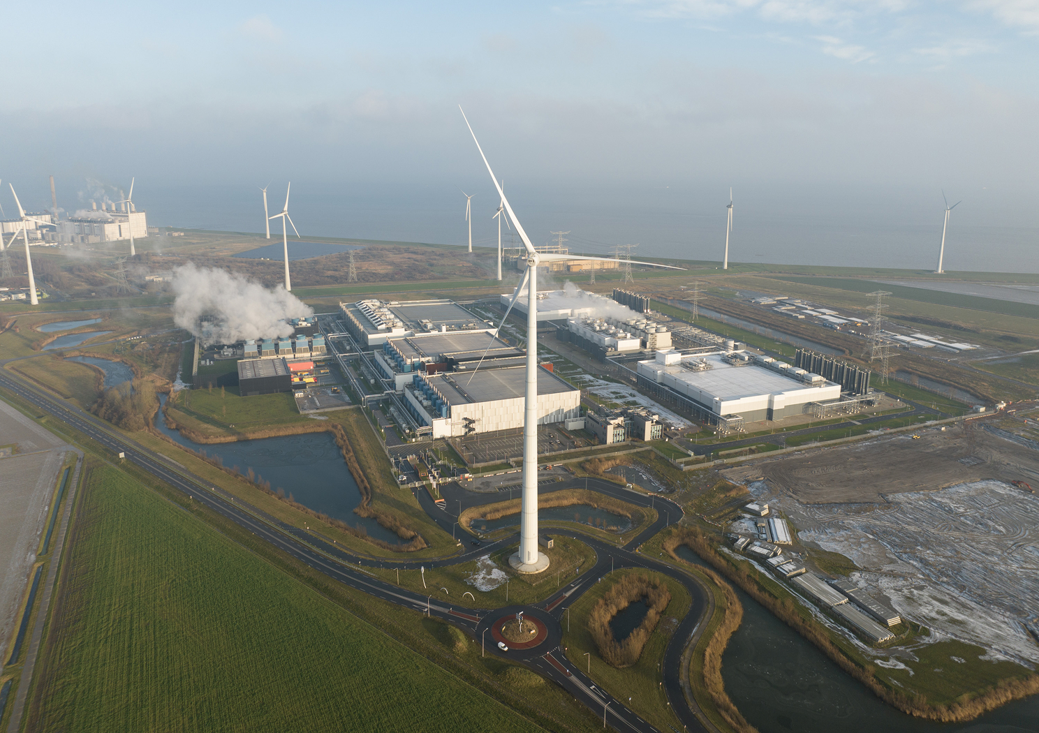Coastal power facility with wind turbines and industrial structures beside the sea.