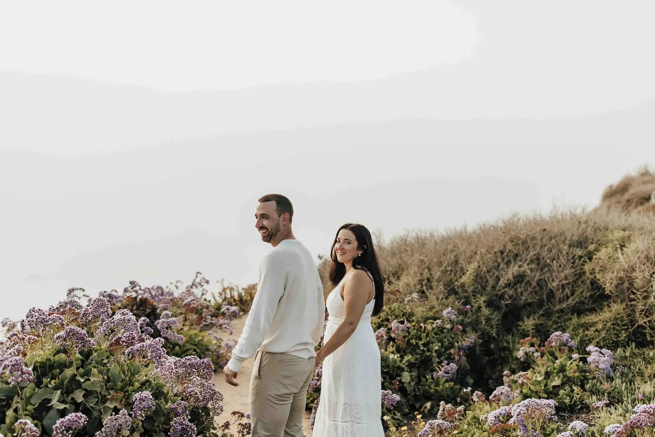 Engaged couple walking hand in hand along a flower-covered coastal cliff overlooking the ocean in San Diego.