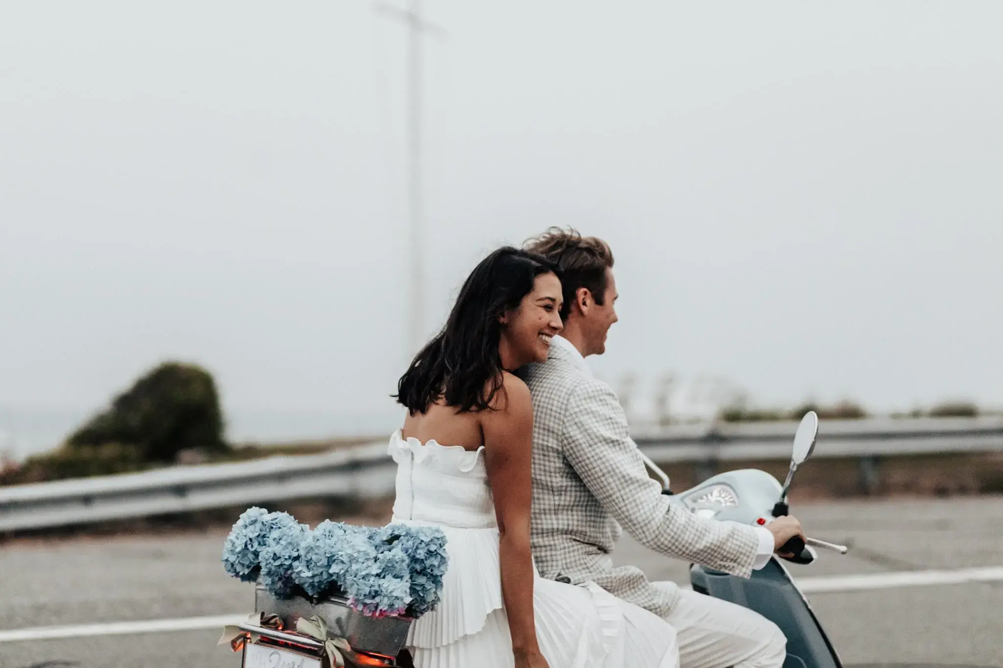 Bride and groom riding a vintage scooter after their Little Compton, Rhode Island elopement, with blue hydrangeas tucked into the back and the coastal road ahead.