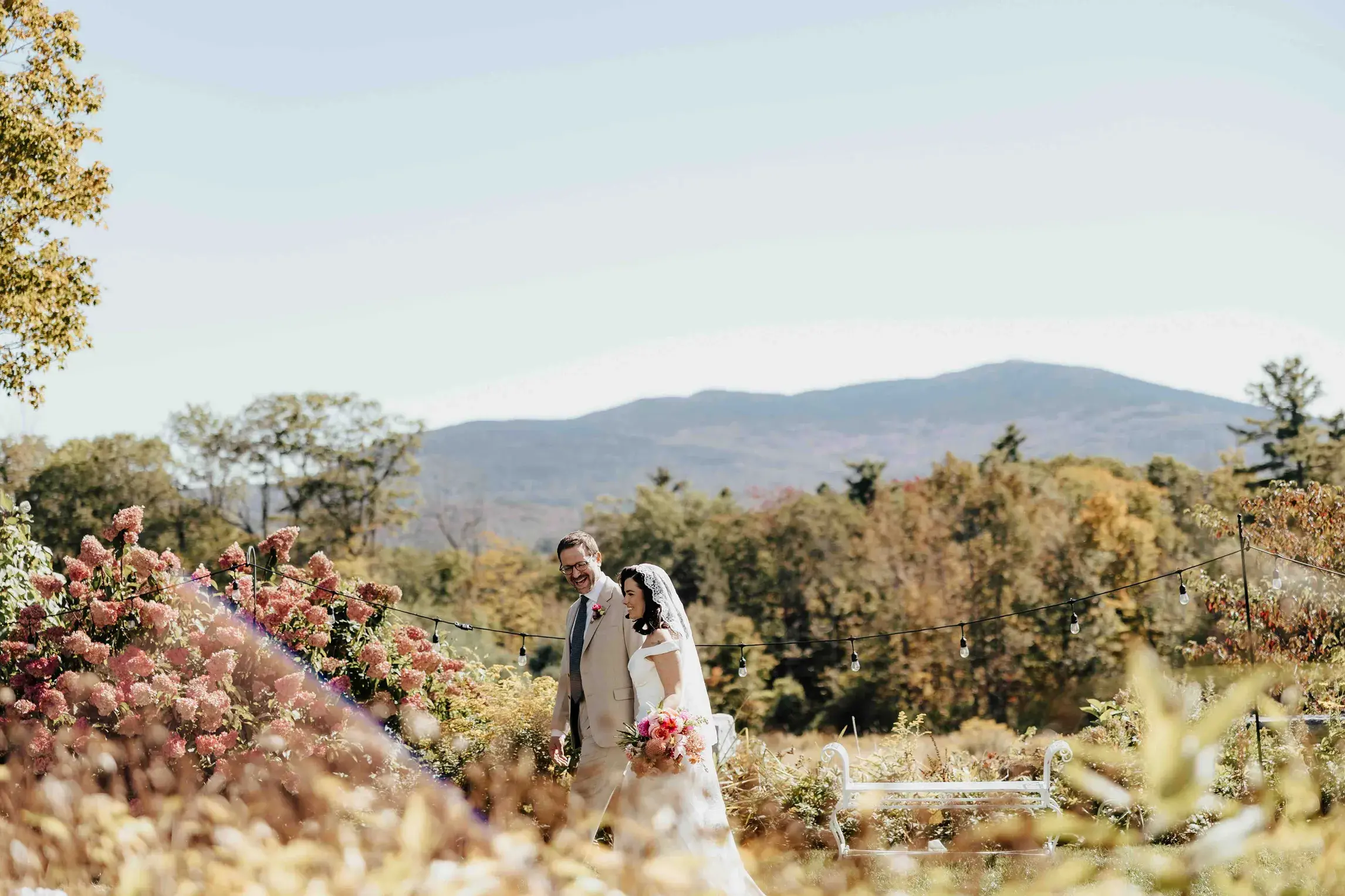 Bride and groom strolling through the gardens at Aldworth Manor during their wedding in Harrisville, New Hampshire, with colorful blooms and mountain views in the background.
