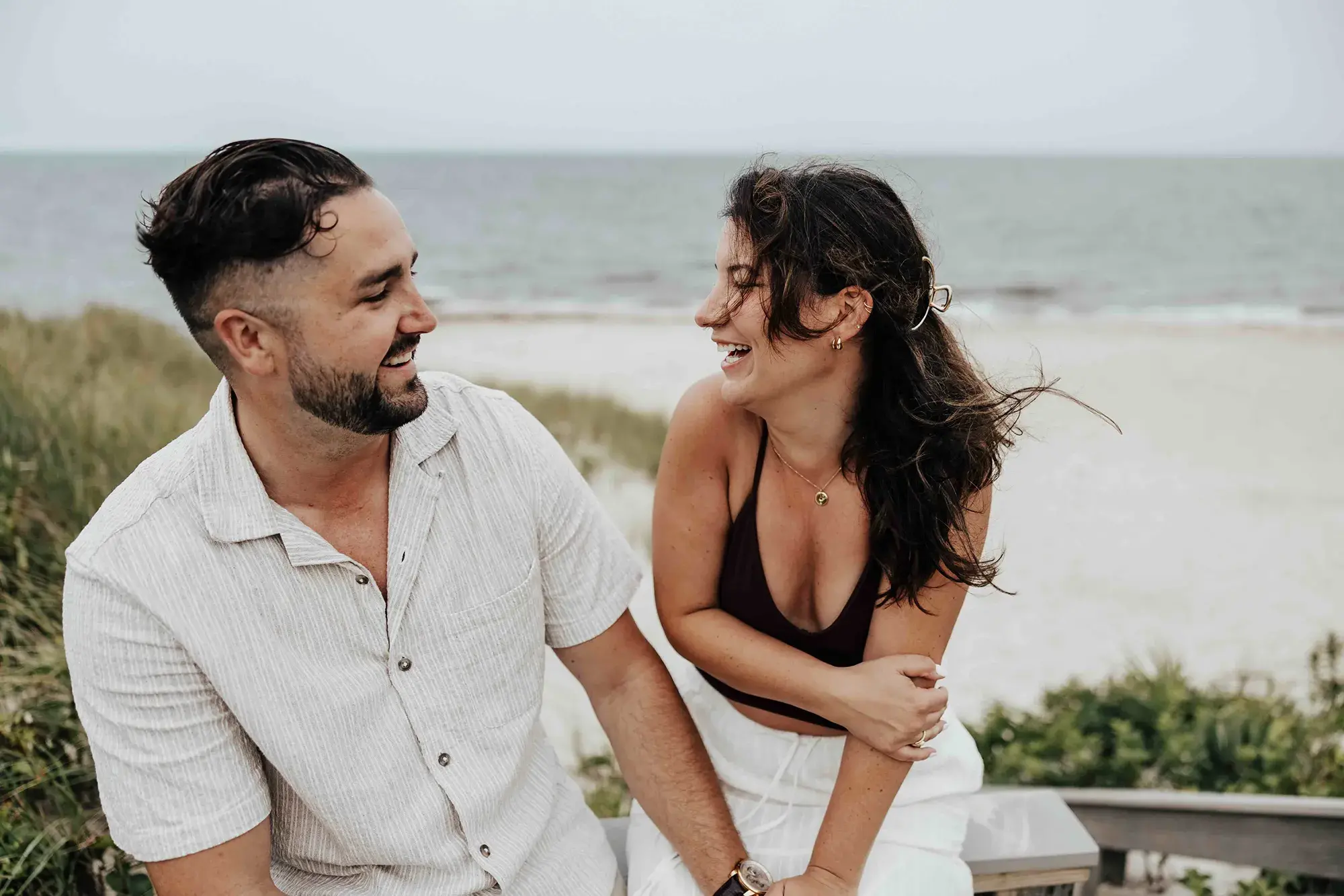 Couple laughing together by the beach after a Cape Cod proposal, captured during a relaxed coastal session with the ocean in the background.