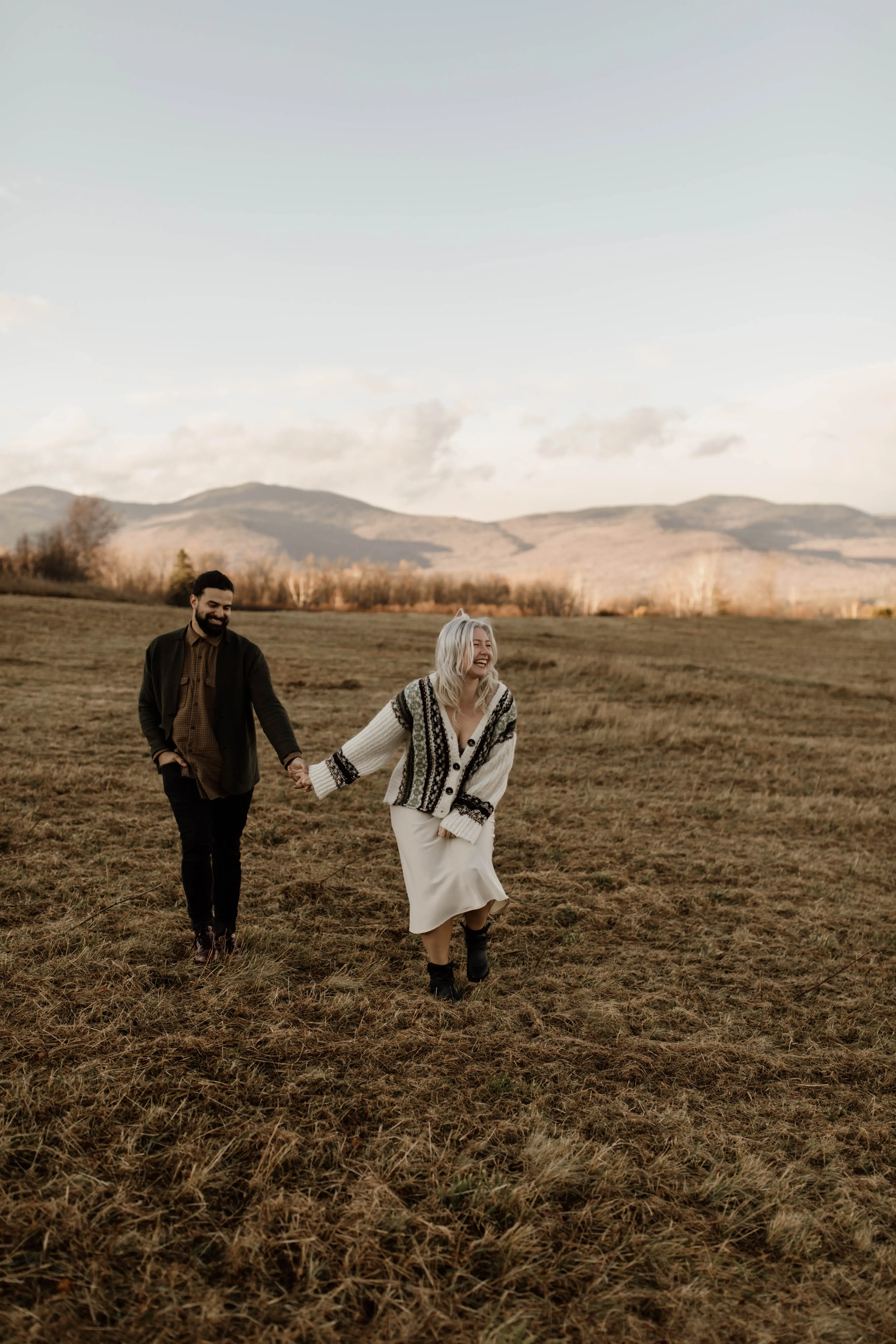 New England photographer walking hand in hand with her partner through an open field, with rolling mountain views stretching across the background.
