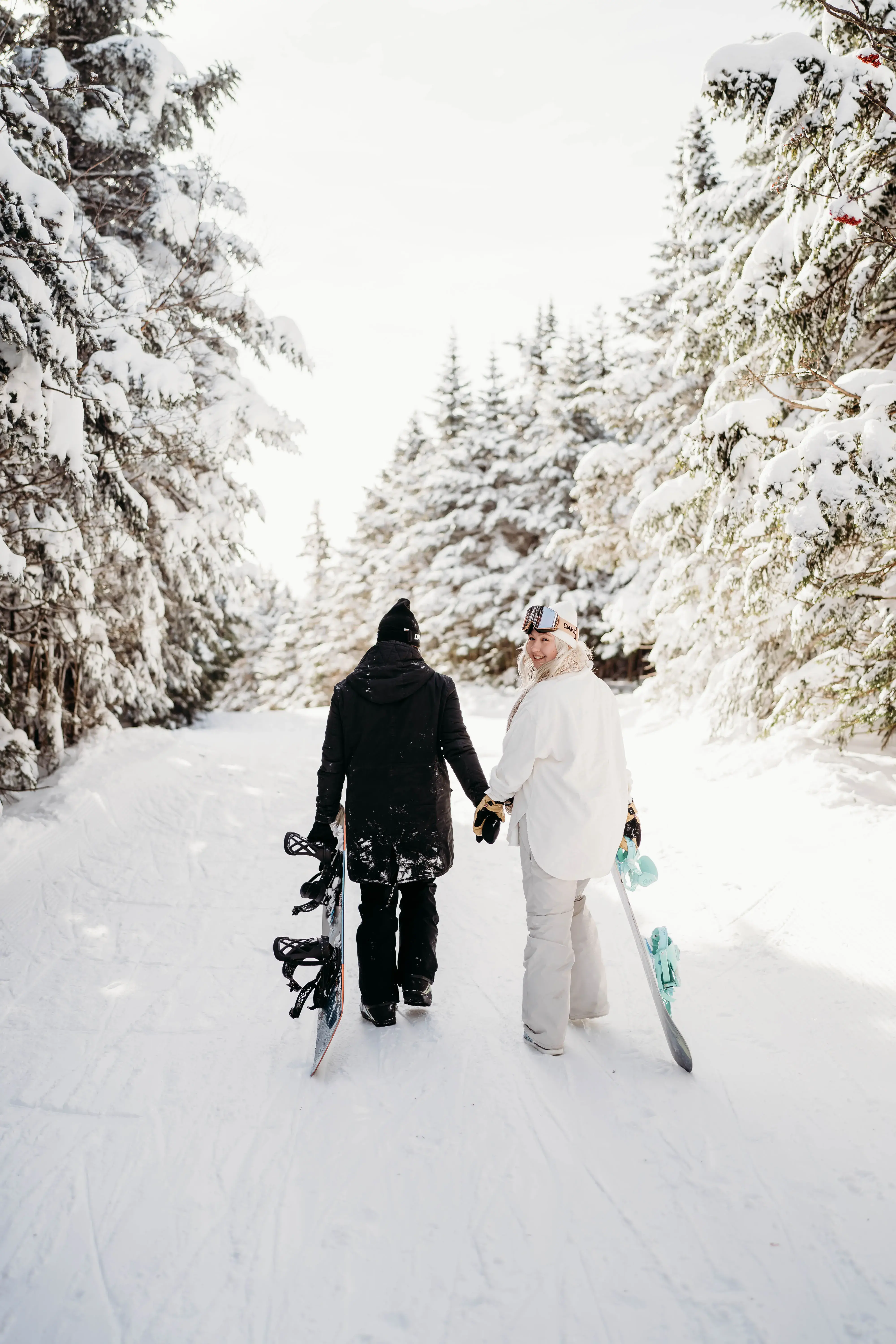Vermont wedding photographer walking hand in hand with a partner through a snow-covered forest while carrying snowboards after a winter day on the mountain.