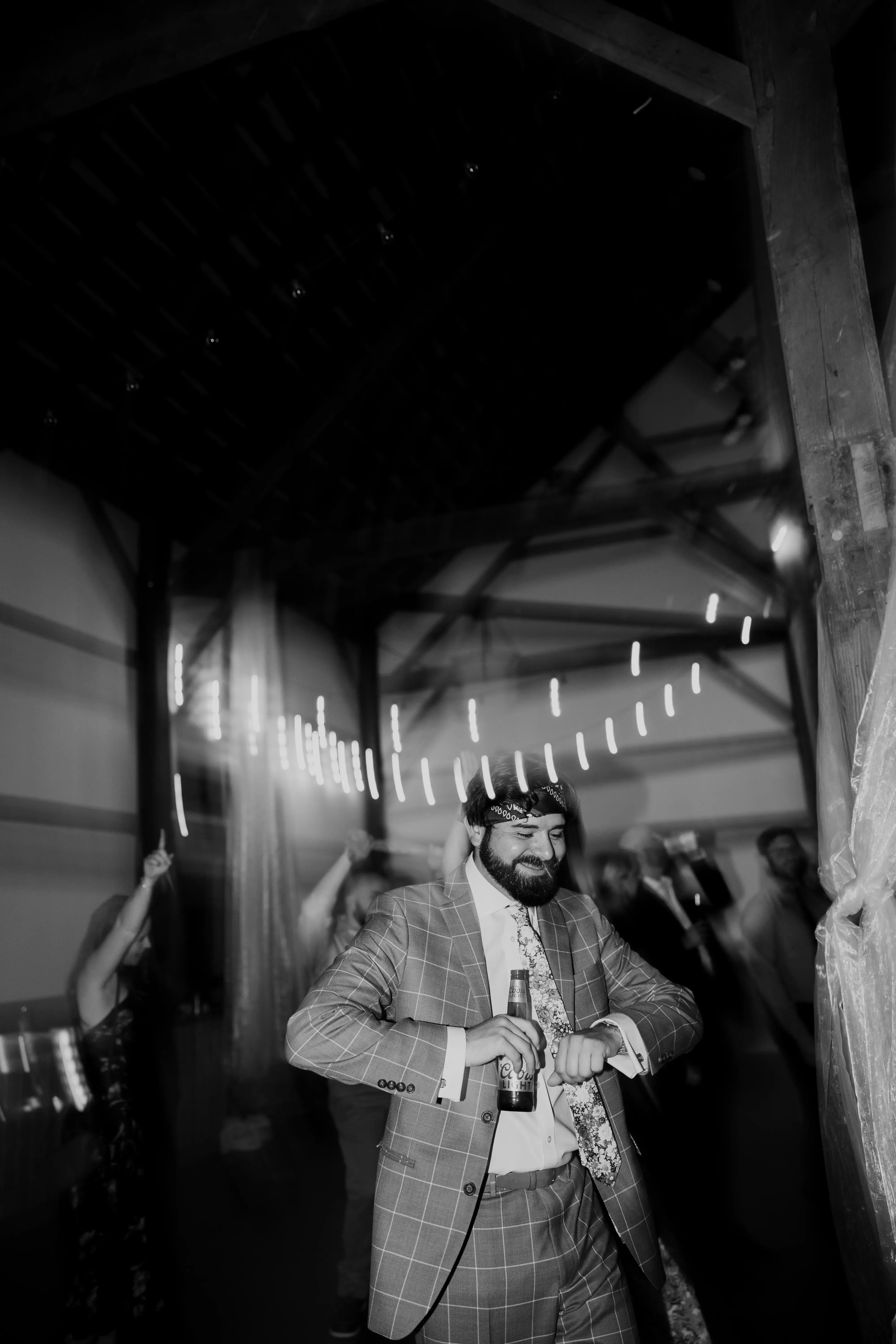 Groom dancing at the wedding reception in a checkered suit and bandana, holding a beer beneath string lights as guests celebrate around him.