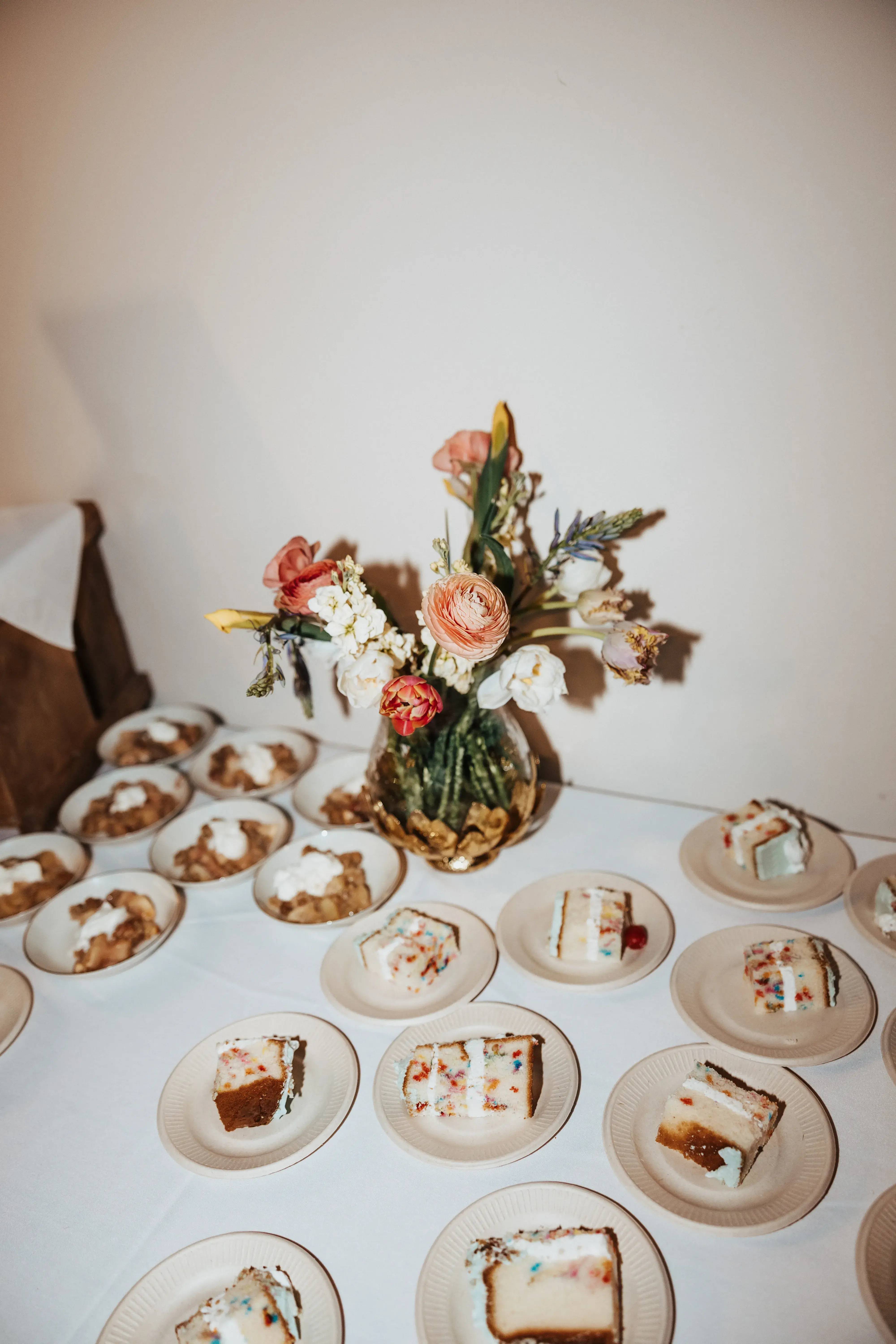 Slices of wedding cake and dessert bowls arranged on a reception table beside a colorful floral centerpiece in a glass vase.