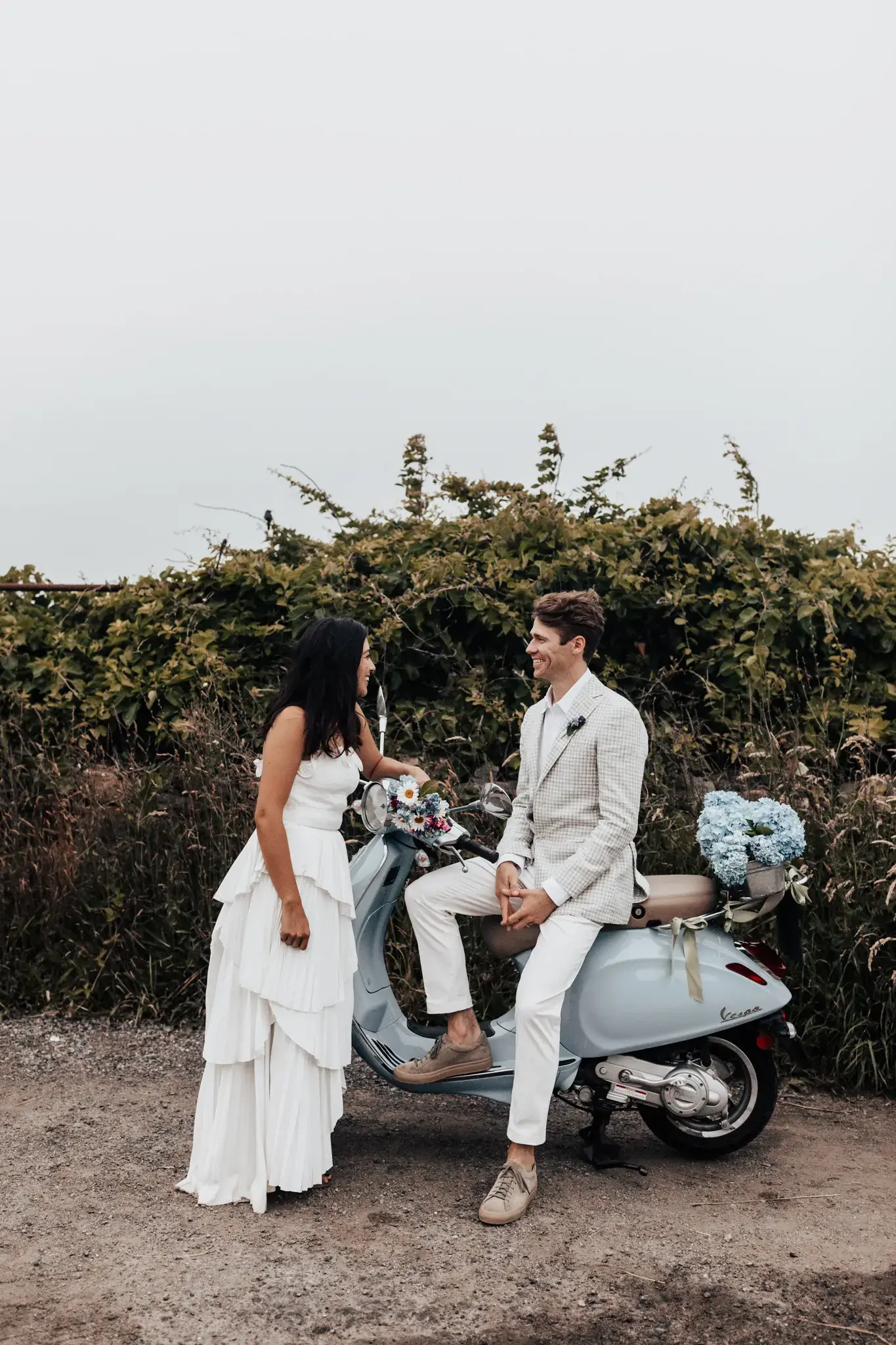 Bride and groom sharing a relaxed moment beside a vintage Vespa decorated with flowers during their outdoor wedding celebration.