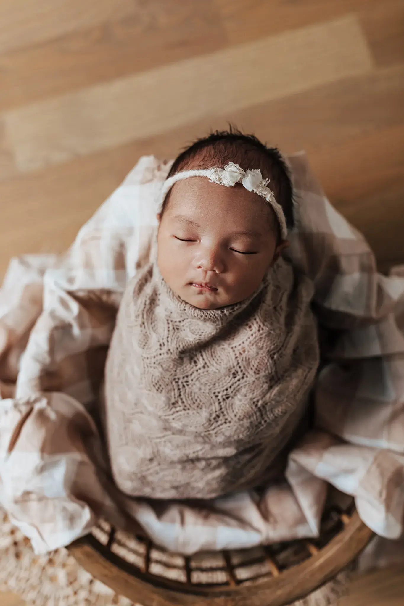 Newborn baby sleeping peacefully while wrapped in a soft lace blanket and floral headband during a warm, intimate portrait session.