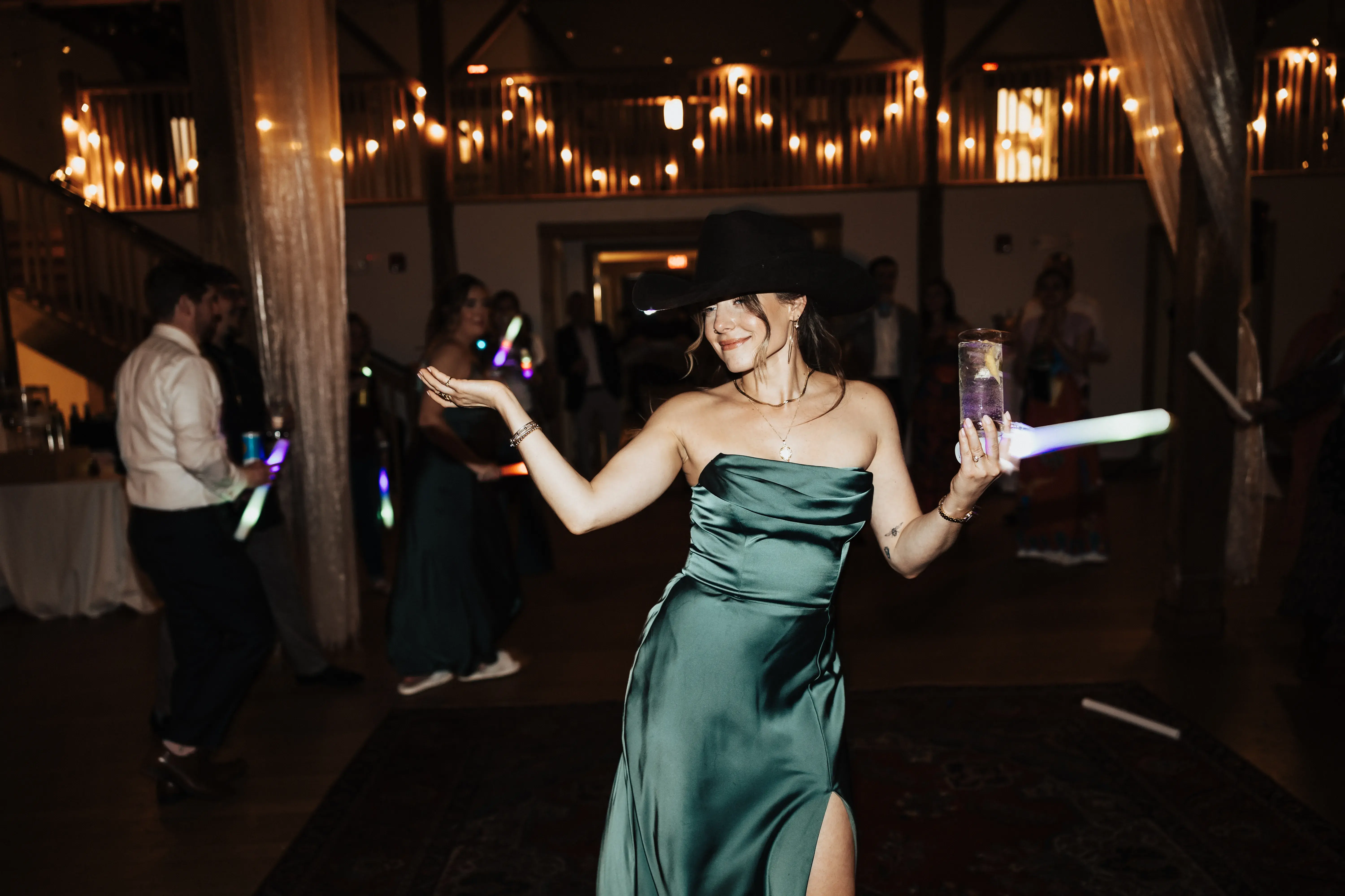 Wedding guest dancing in a green satin dress and black cowboy hat while holding a drink and glow stick beneath warm string lights at the reception.