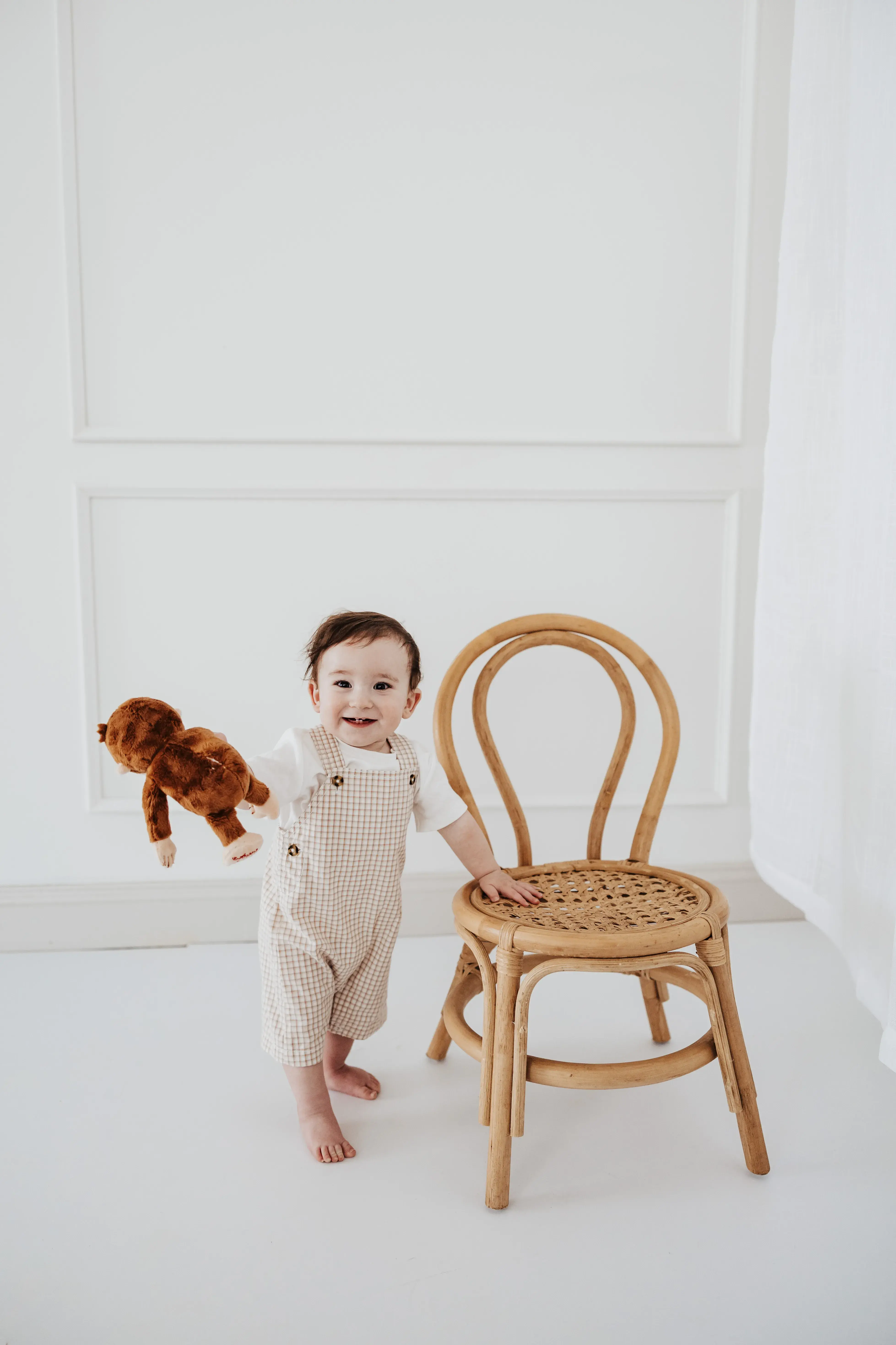 Smiling toddler holding a stuffed monkey while standing beside a wooden chair during a bright and simple studio portrait session.