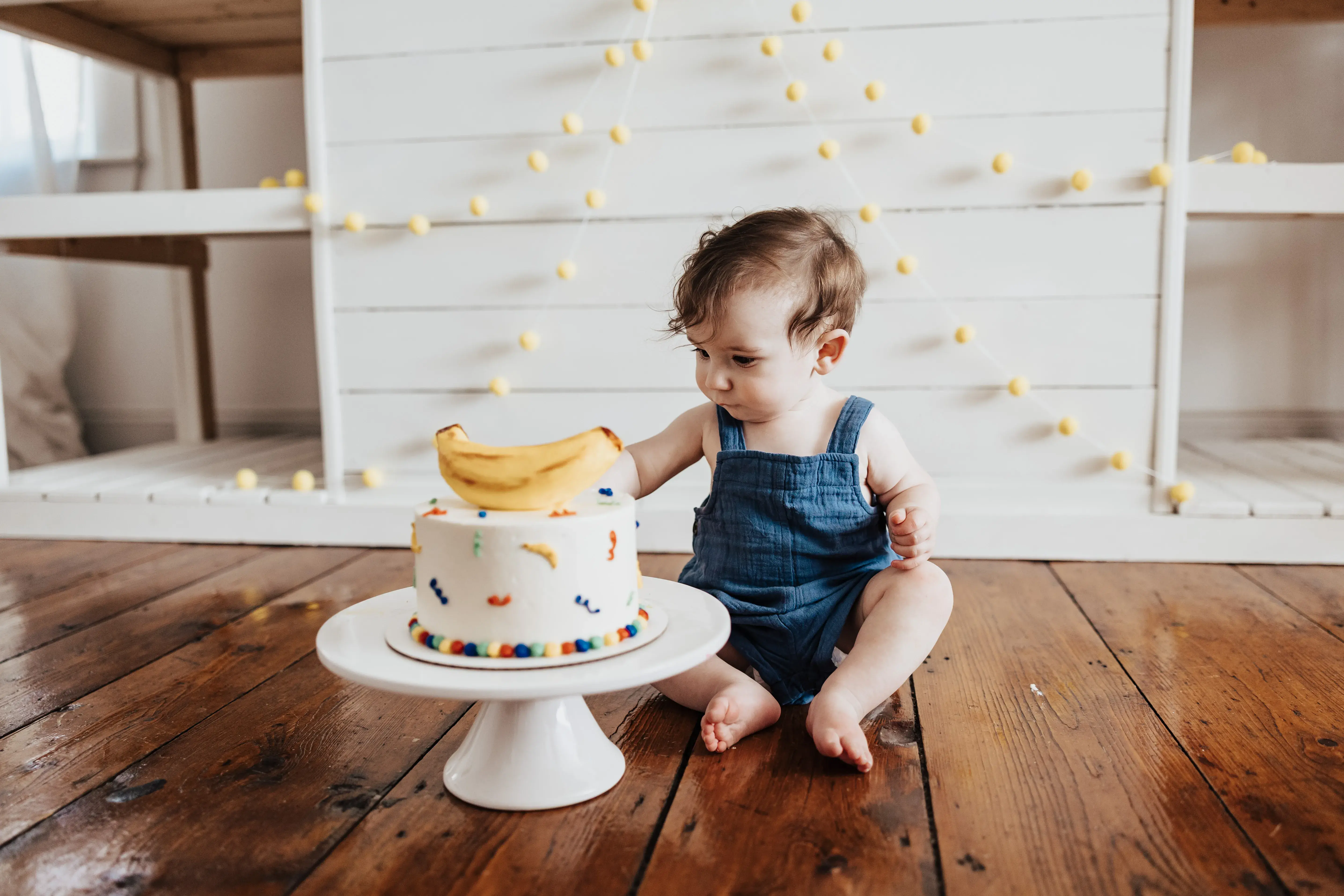 Baby reaching toward a colorful birthday cake topped with bananas during a playful first birthday cake smash session on a wooden floor.