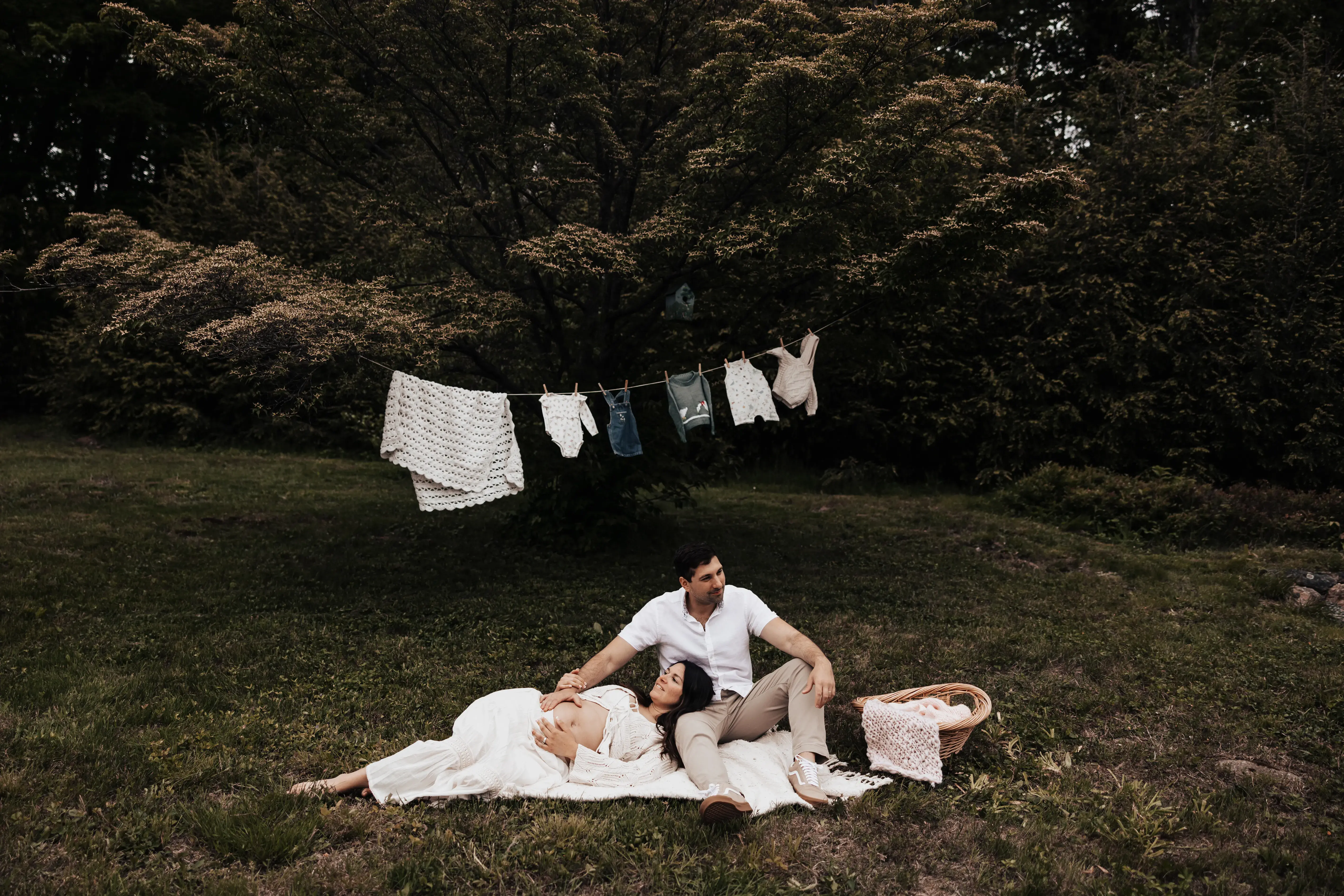 Expectant parents relaxing together on a blanket in a grassy field during a maternity session, with baby clothes and a handmade blanket hanging on a clothesline behind them.