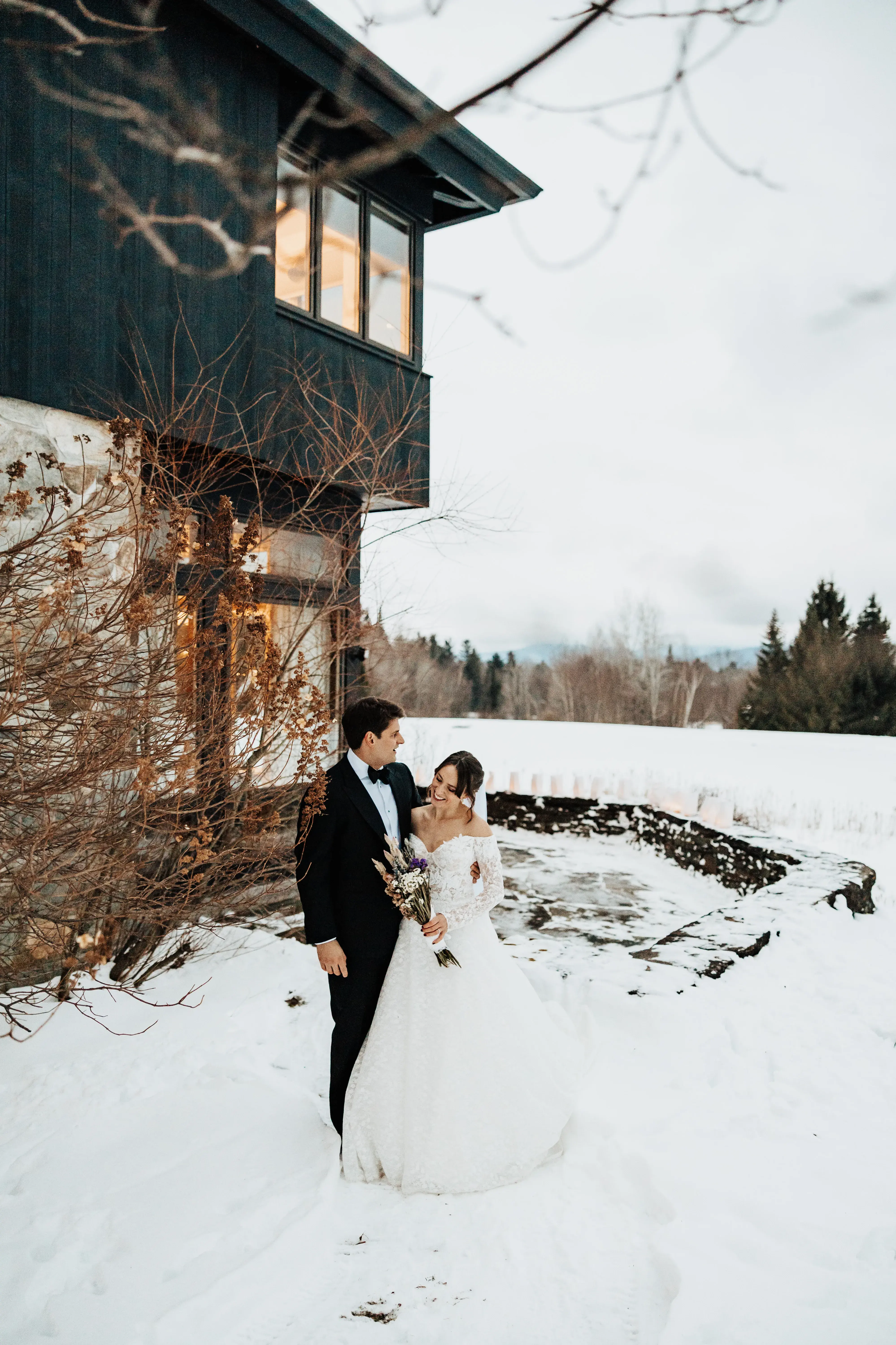 Bride and groom smiling together outside a modern lodge during a snowy winter wedding, surrounded by fresh snow and quiet mountain scenery.