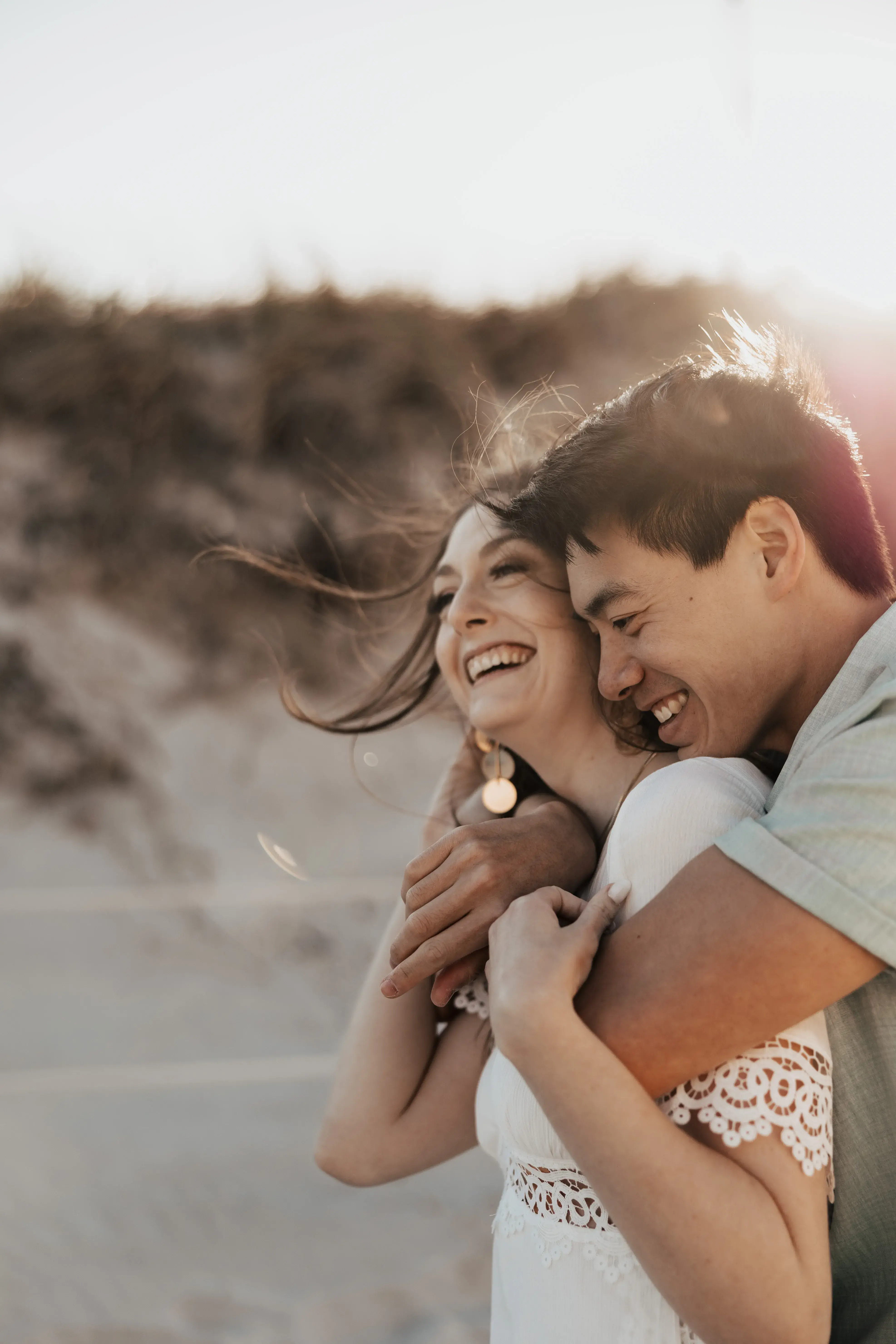 Couple laughing and embracing on a sunlit beach, sharing a warm and playful moment with soft golden light behind them.