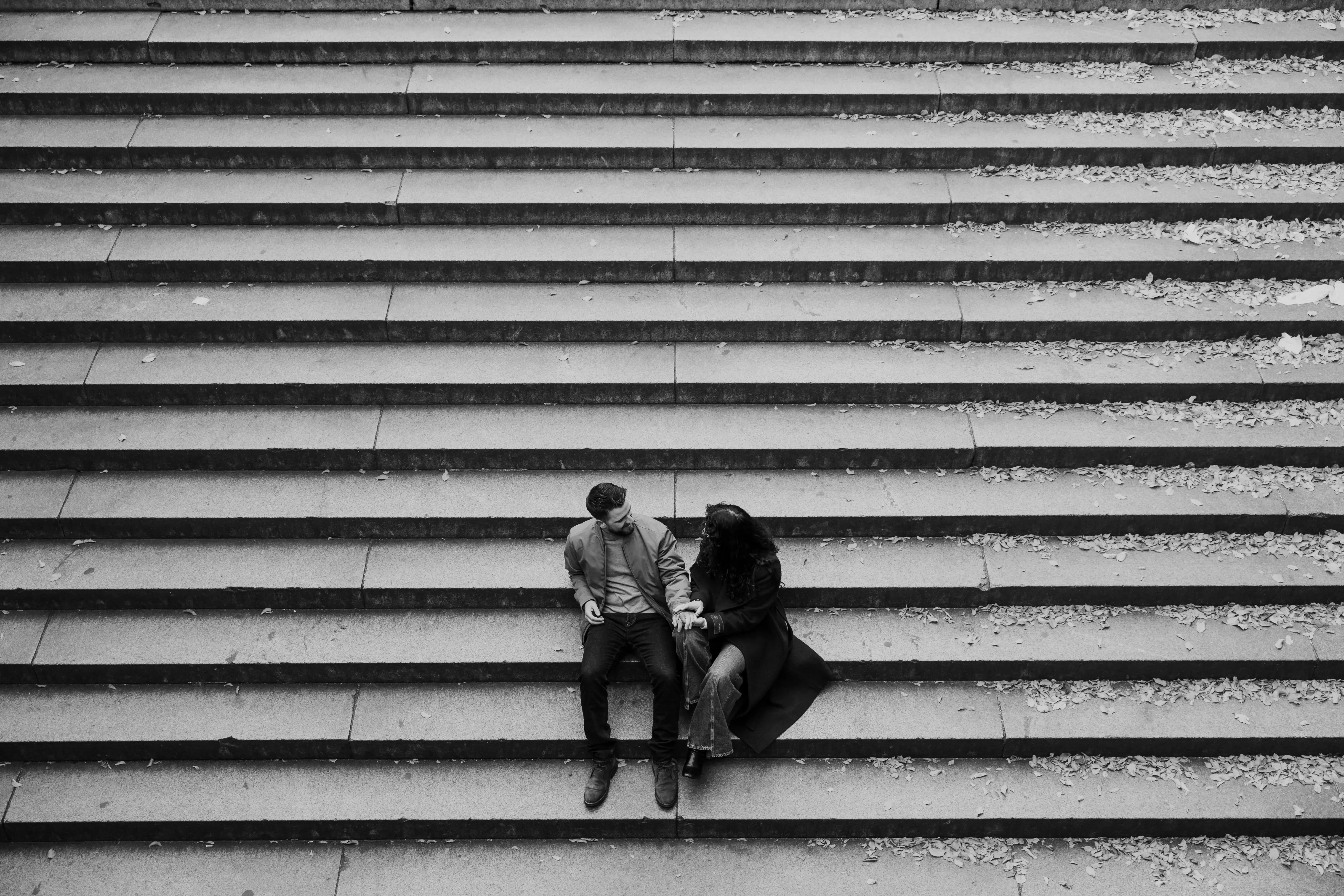Couple sitting close together and holding hands on wide stone steps, captured in a candid black-and-white engagement portrait.